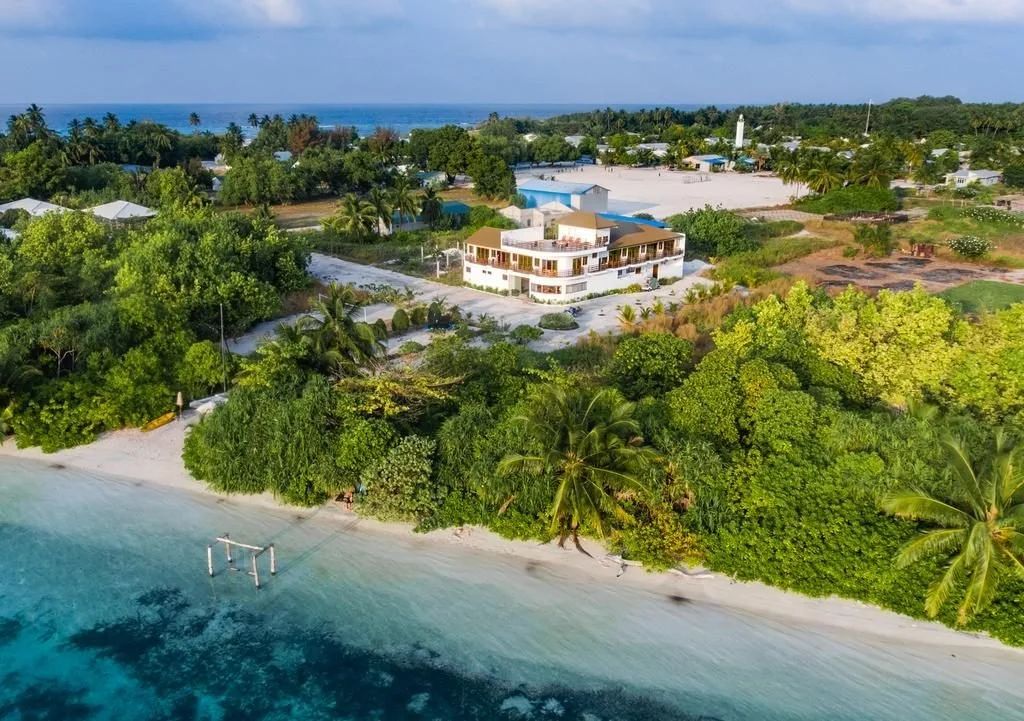 Aerial view of a tropical beach with white sand, lush green trees, and a large white building near the shoreline, overlooking turquoise ocean waters with a volleyball net set up on the beach.