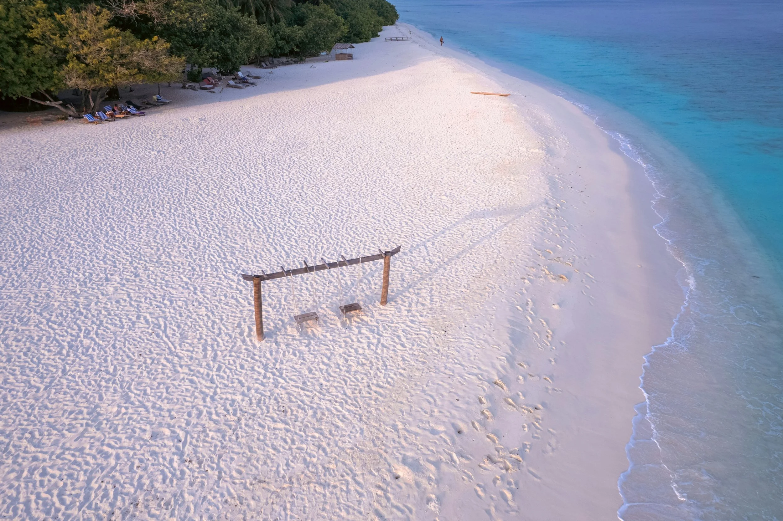 Empty sandy beach with a wooden swing set, shoreline with gentle waves, and green trees in the background.
