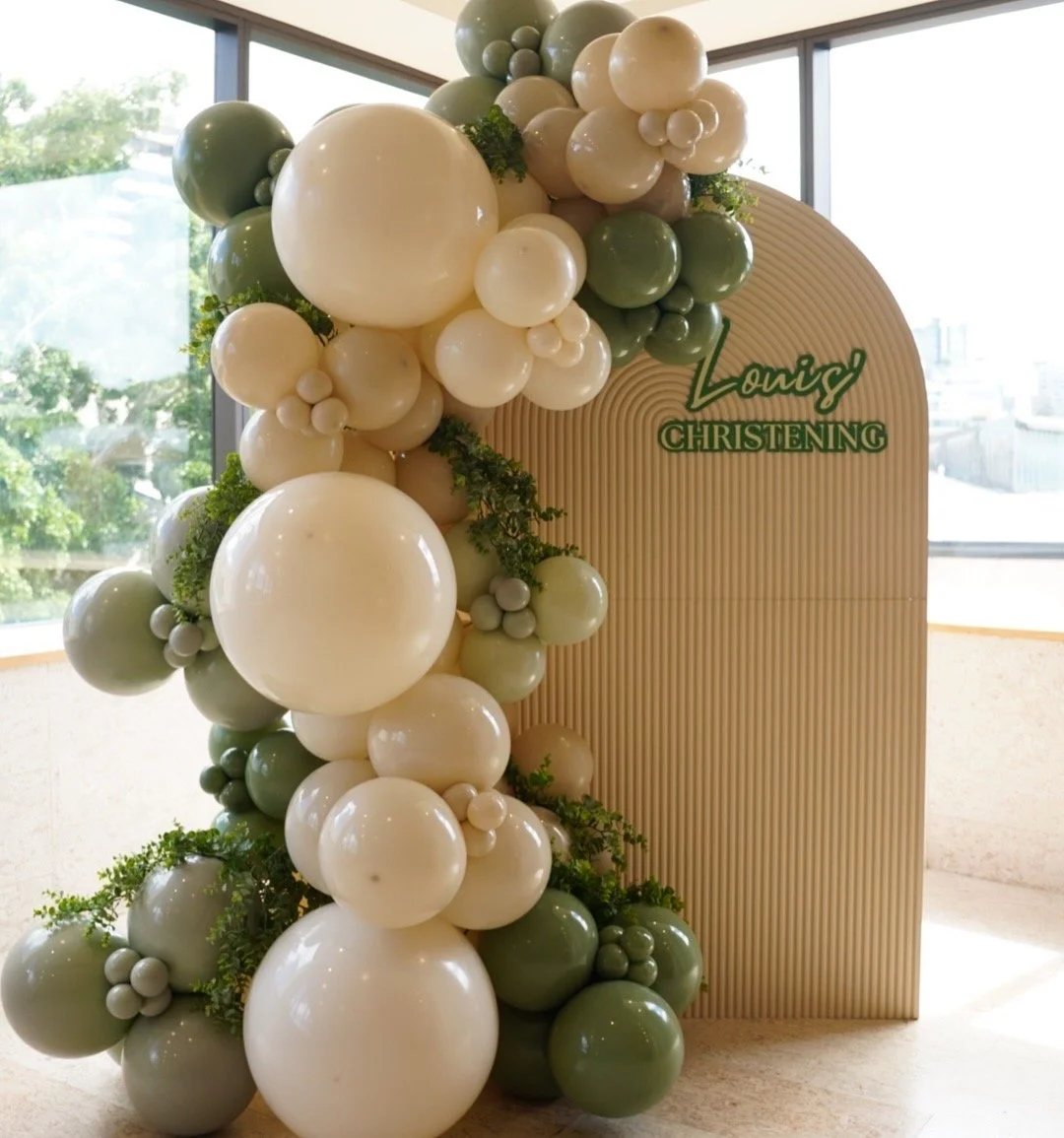 A decorative balloon arrangement with white, cream, and green balloons, along with greenery, next to a beige backdrop with green text that reads 'Loning Christening'.