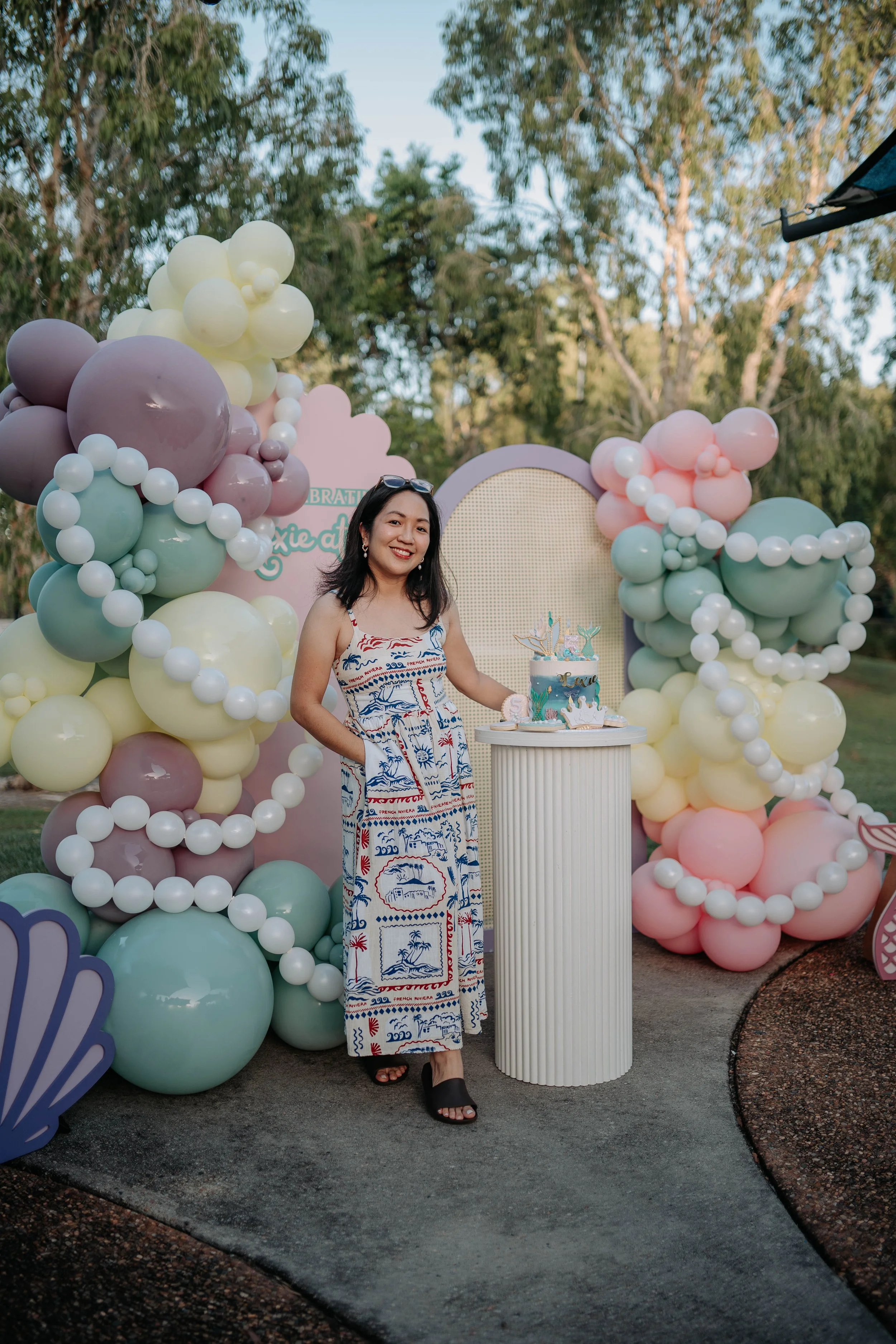 A woman in a sleeveless dress with a tropical pattern stands outdoors near a celebration setup with pastel balloon decorations and a cake, smiling at the camera.