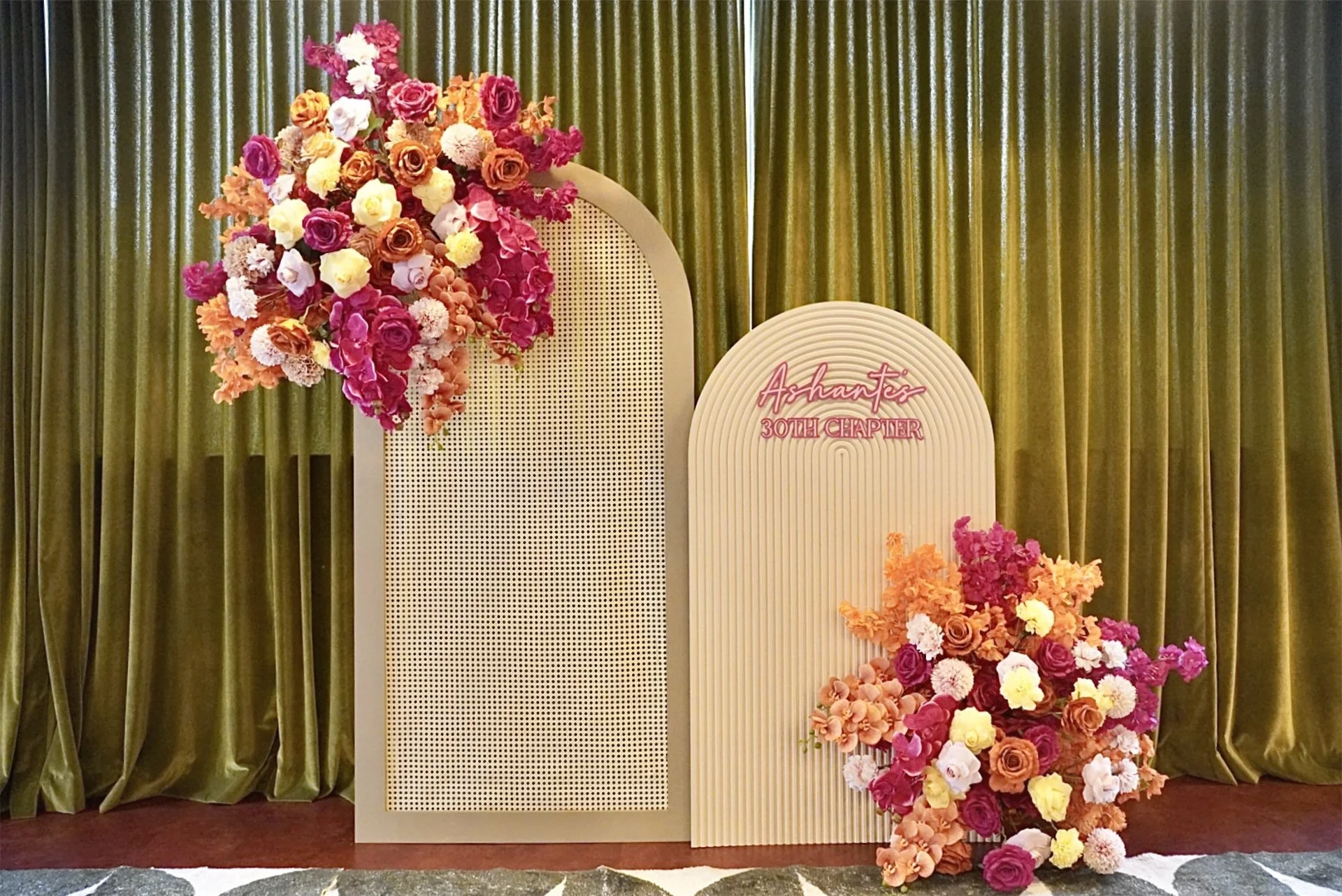 Floral display with pink, orange, white, and purple flowers on a white backdrop with green curtains in the background and a pink neon sign reading 'Ashanti 30th Chapter'.