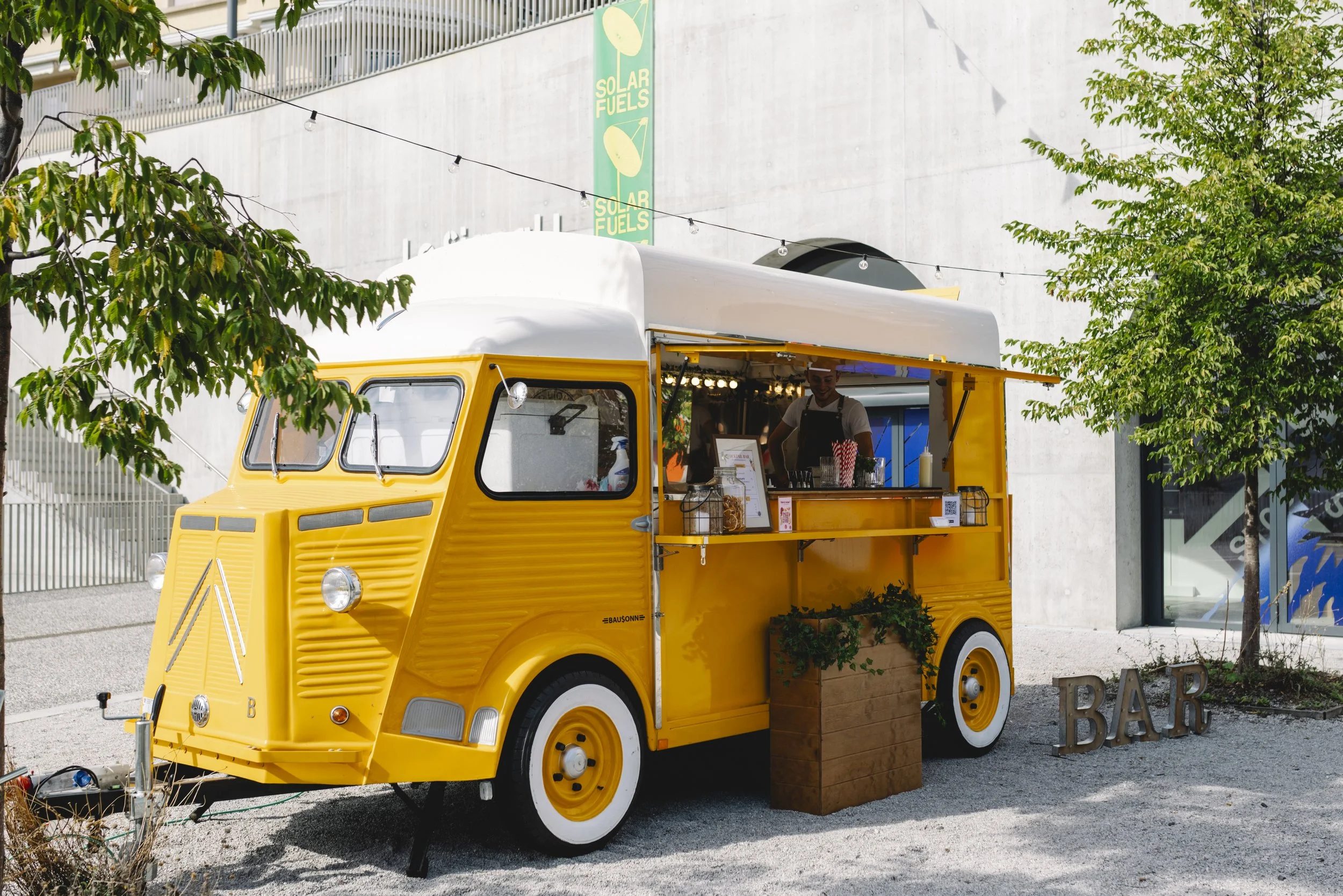 Un camion de nourriture jaune converti en bar, avec un comptoir ouvert et un employé à l'intérieur, situé en plein air sous un arbre. La scène inclut des plantes en pot et une enseigne indiquant 'BAR' en fer dehors.
