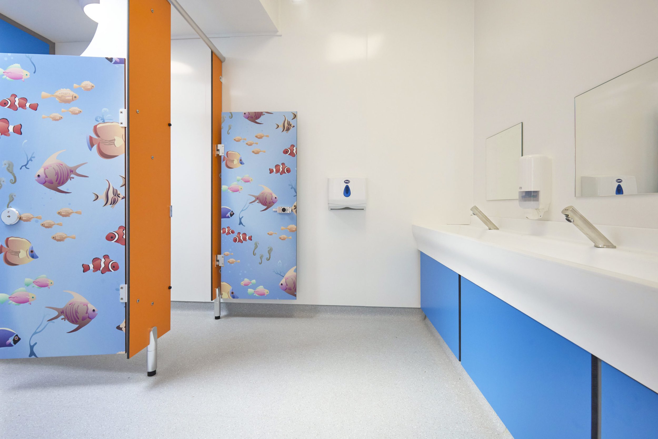 a blue vanity unit and solid surface wash trough with touch free taps, child lower cubicles in orange and waterworld print in a washroom at upland school.jpg