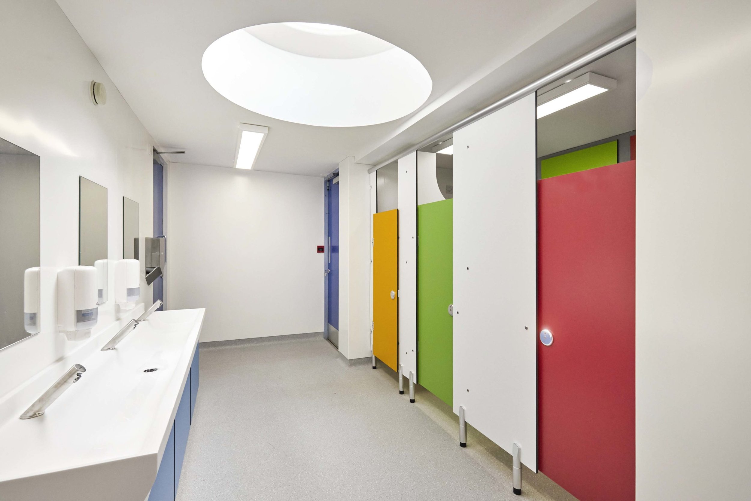 a washroom with a vanity unit in blue with a hand wash trough on top, mirrors, white hygienic cladding on walls, and lower child cubicles with multi coloured doors at upland school.jpg