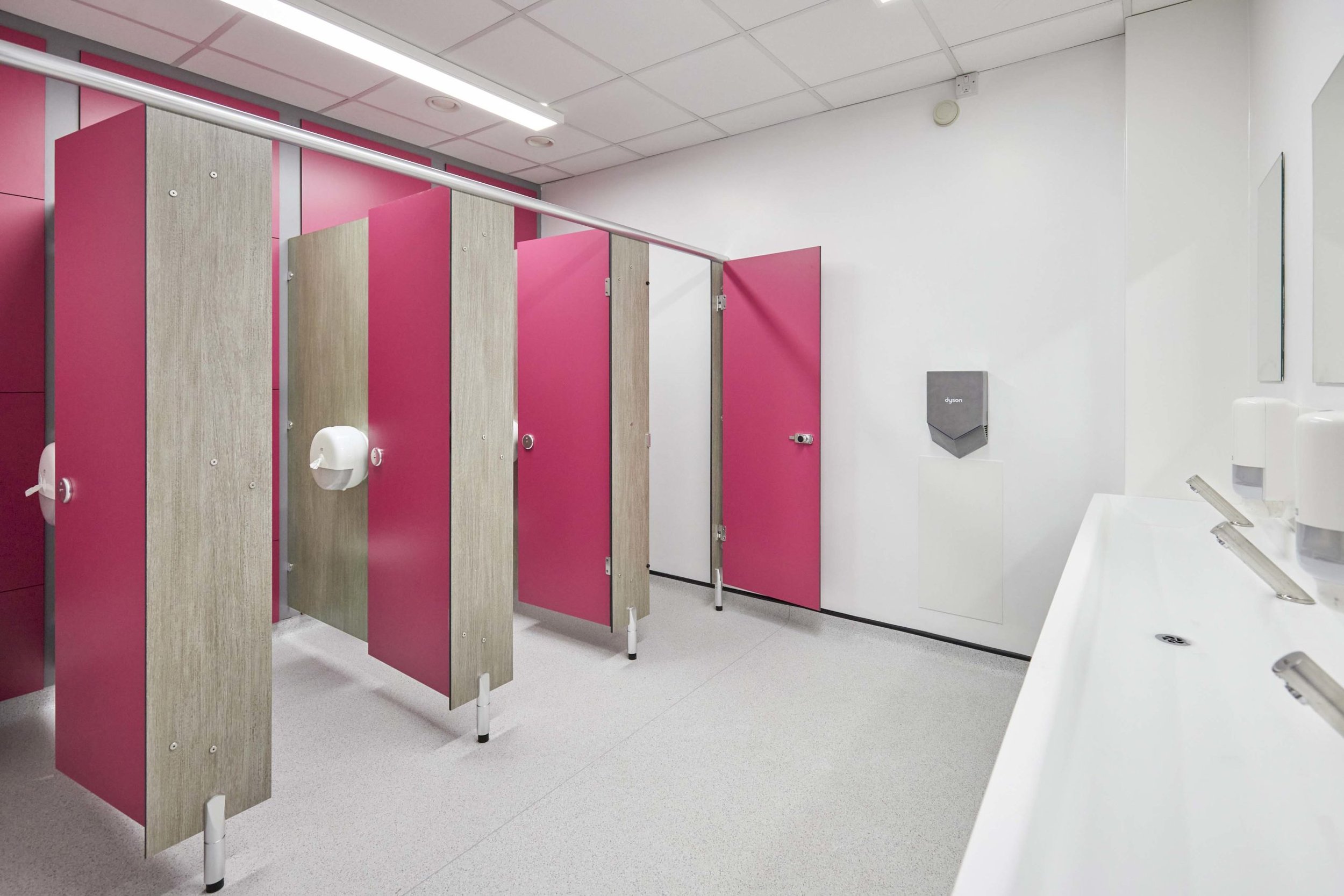 a washroom with white walls, a dyson hand dryer, a solid surface vanity trough with touch free taps, pink duct panels, cubicles in grey woodgrain with pink doors at upland school.jpg