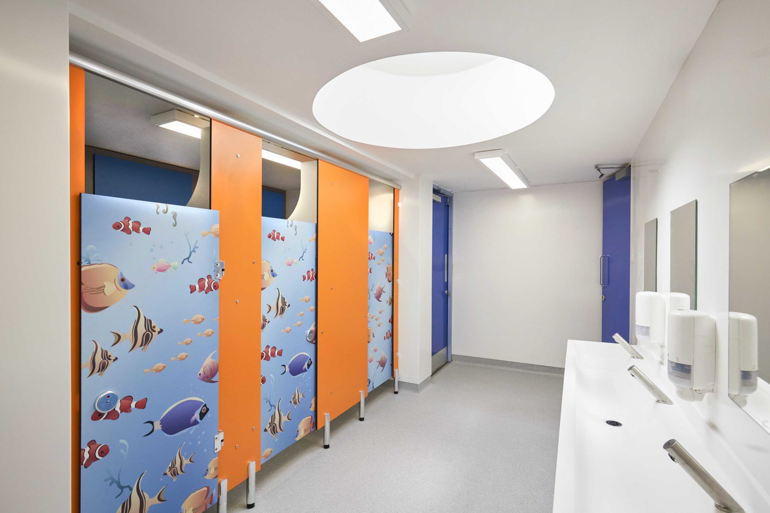 washroom with blue entrance doors, a vanity unit with a solid surface wash trough top and mirrors above, and child lower cubicles with orange pilasters and waterworld pattern doors.jpg