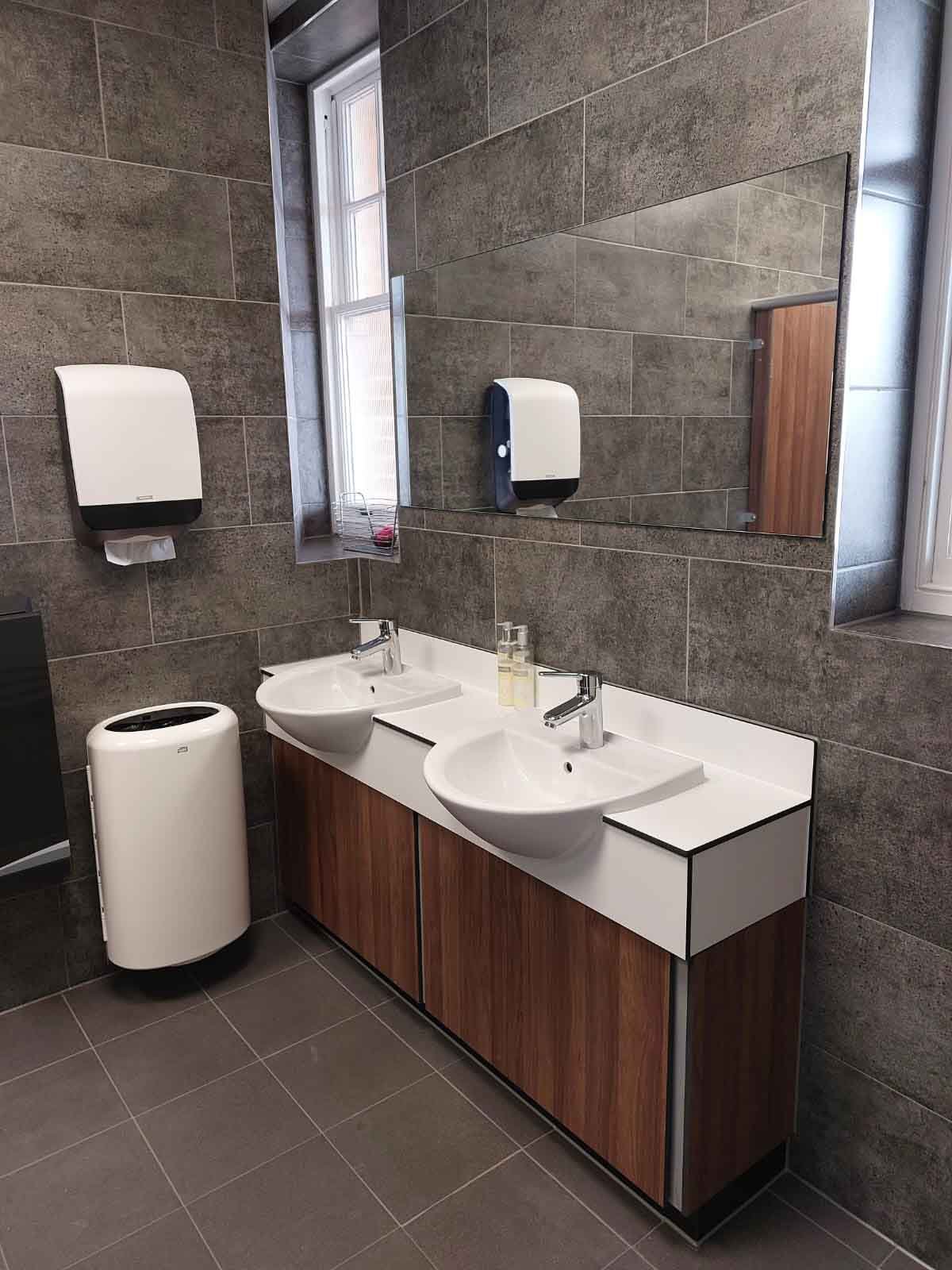 washroom with grey tiling, a hand wash station with bin and paper dispenser, a woodgrain vanity unit with a white vanity top and sinks with a mirror above at kia oval.jpg