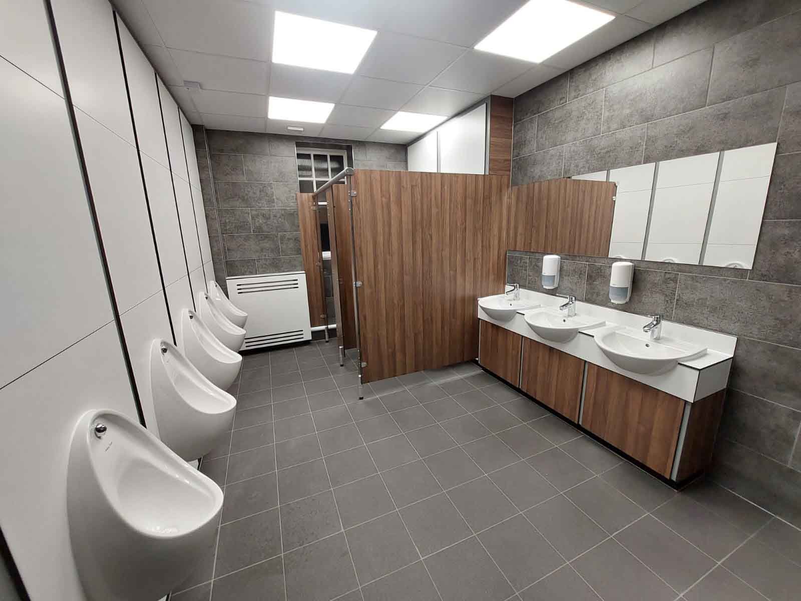 a washroom at kia oval with grey tiling, white duct panelling, woodgrain cubicles and vanity with a white top, a white radiator cover and a mirror.jpg