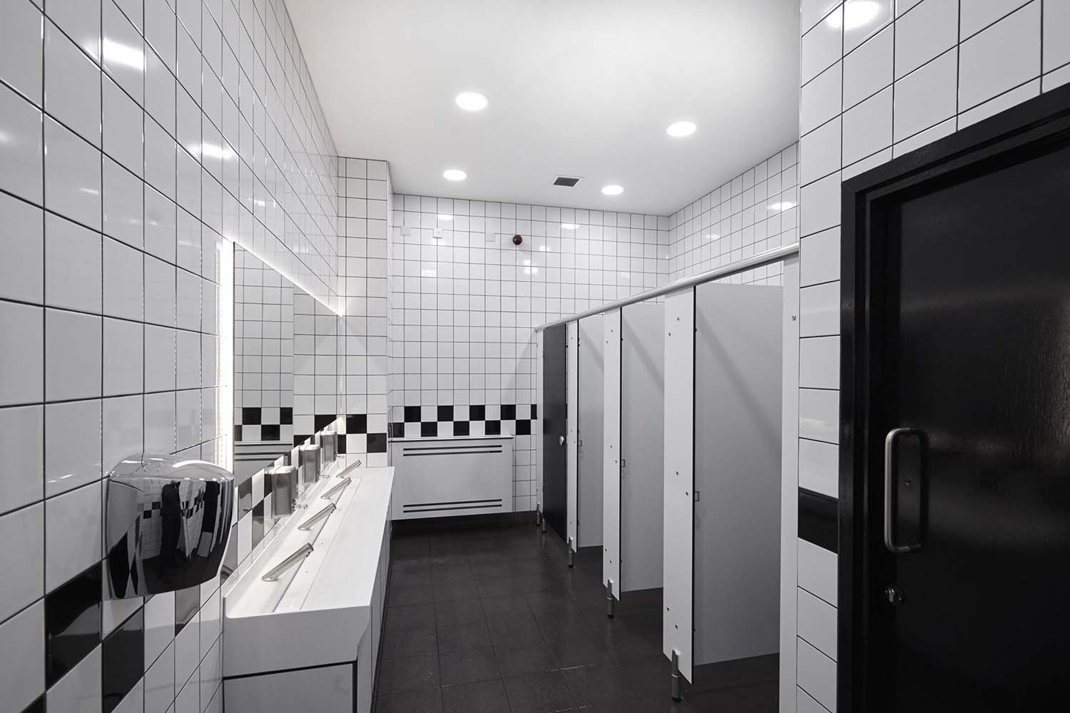 washroom at coventry bus station with black and white tiling, cubicles and a vanity unit with solid surface trough and touch free taps and a backlit mirror above.jpg