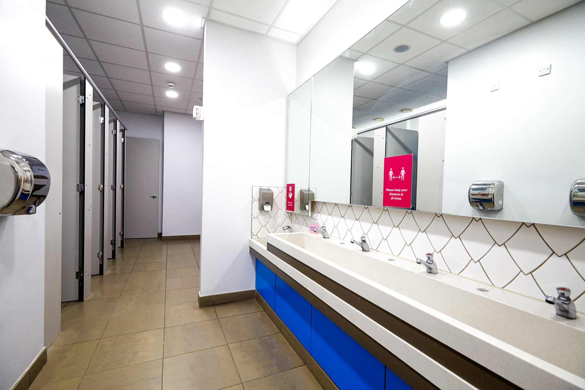 washroom with a feature tile wall, toilet cubicles and a blue vanity unit and solid surface hand wash trough with mirror above at national maritime museum.jpg