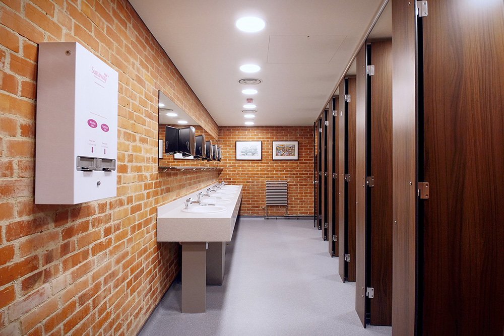 row of woodgrain cubicles and a handwash area, exposed brick walls and a sanitary product dispenser on the wall at glyndebourne opera house.jpg