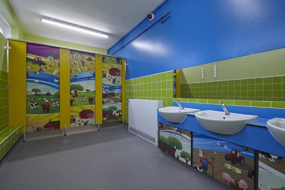 bright childrens washroom with green tiling, blue wall, yellow and farmyard print cubicles and blue vanity unit.jpg