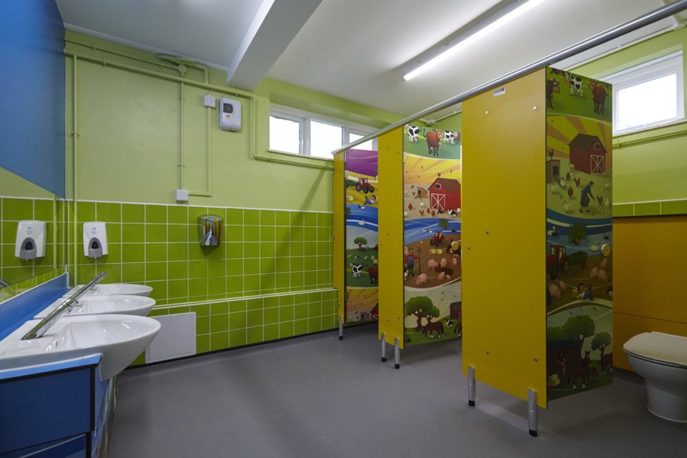 bright childrens washroom with green tiling, yellow and farmyard print cubicles and blue vanity unit.jpg