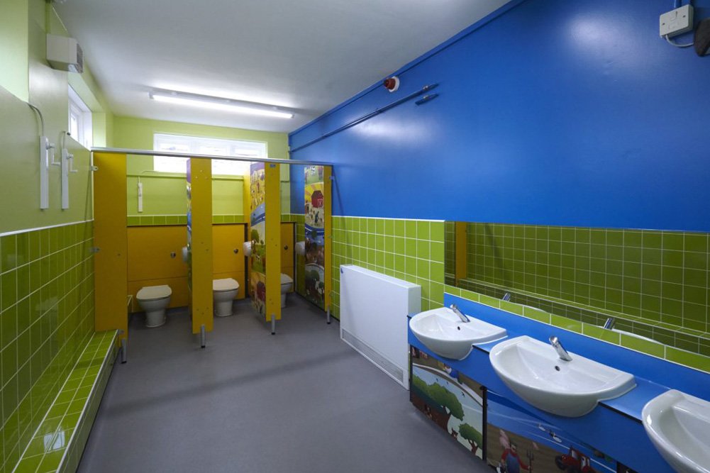 bright childrens washroom with green tiling, yellow and farmyard print toilet cubicles and blue vanity unit.jpg