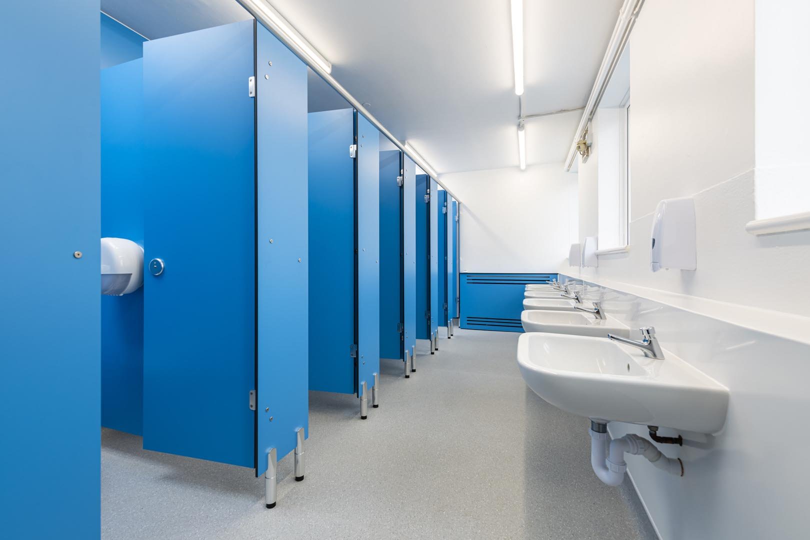 row of blue toilet cubicles and a row of sinks on a wall with hygienic cladding in a washroom at howard junior school.jpg