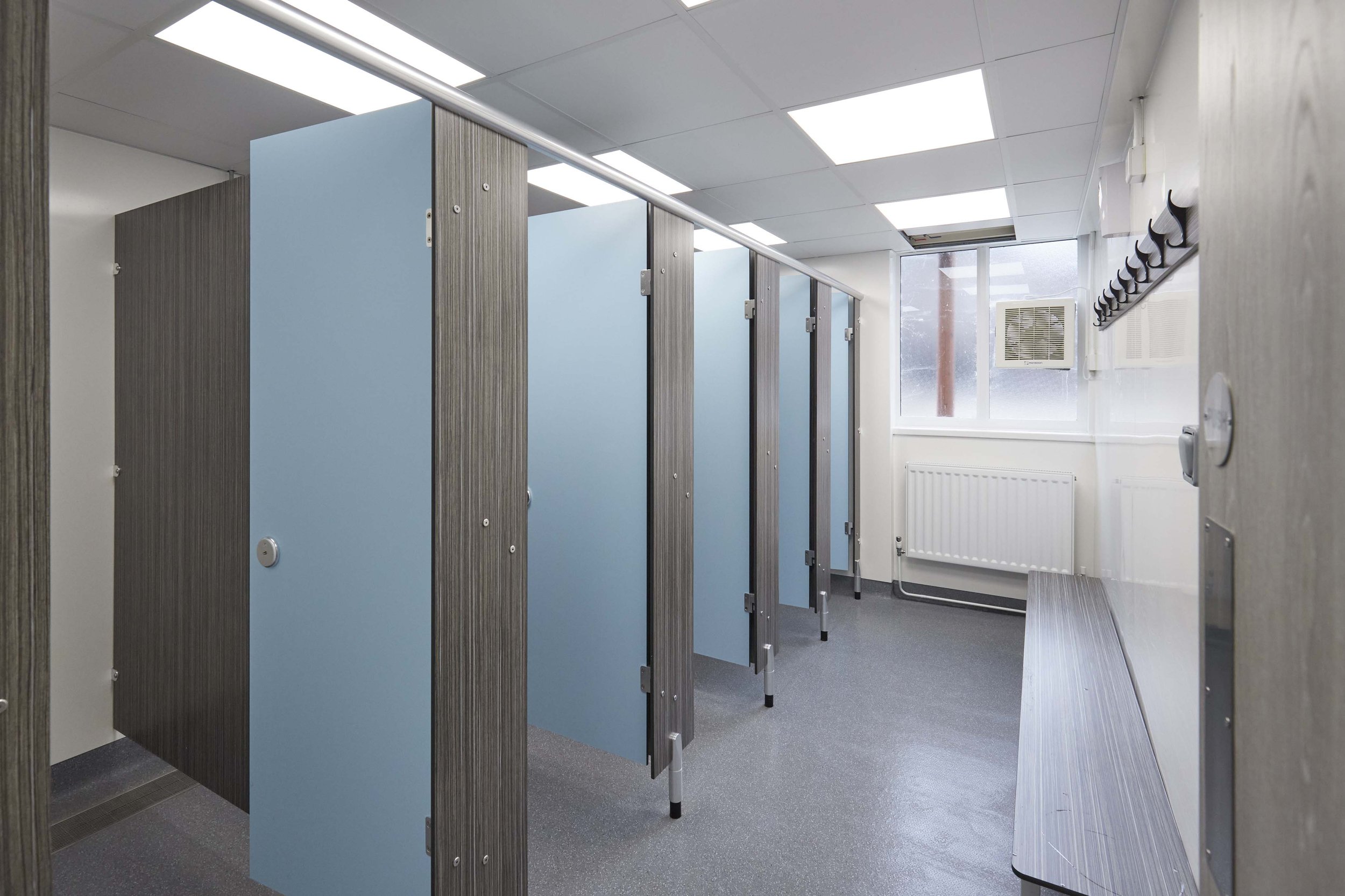 row of shower cubicles in woodgrain and blue colours with hygienic wall cladding on the walls and benching and pegs opposite.jpg