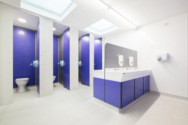 Public restroom with four stalls featuring blue partitions and rolls of toilet paper, and a double sink area with two hands-free soap dispensers and a paper towel dispenser on the wall.
