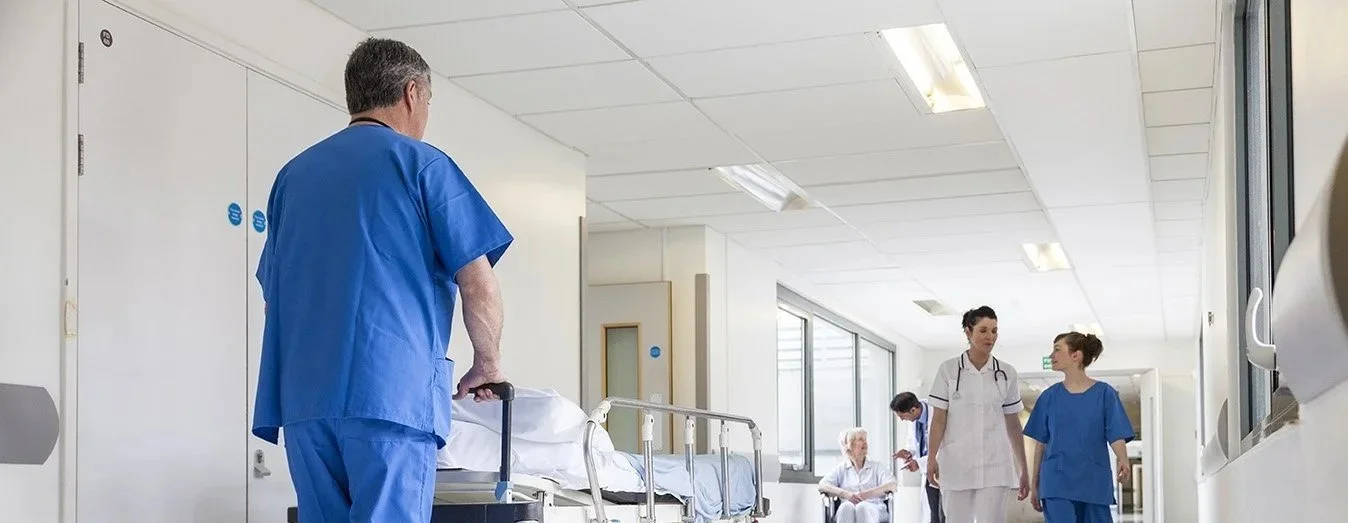 Medical staff, including a male nurse in blue scrubs and female nurses in white and blue scrubs, working in a hospital corridor with a patient bed and large windows.