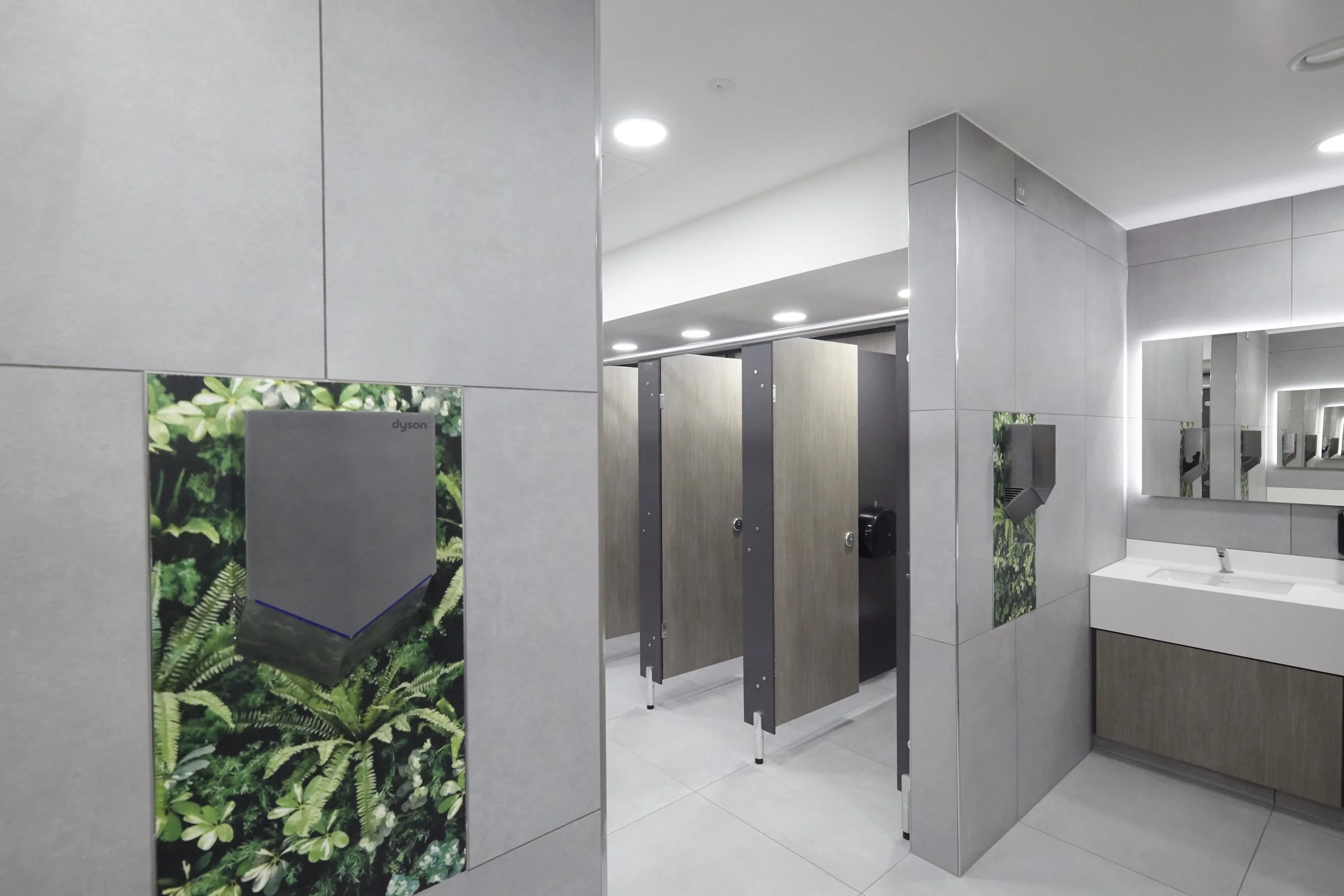 A modern public restroom with gray tiled walls and a row of cubicles. There are back lit mirrors with nature images and a white sink. The ceiling has round recessed lighting, and there are soap dispensers and paper towel holders.
