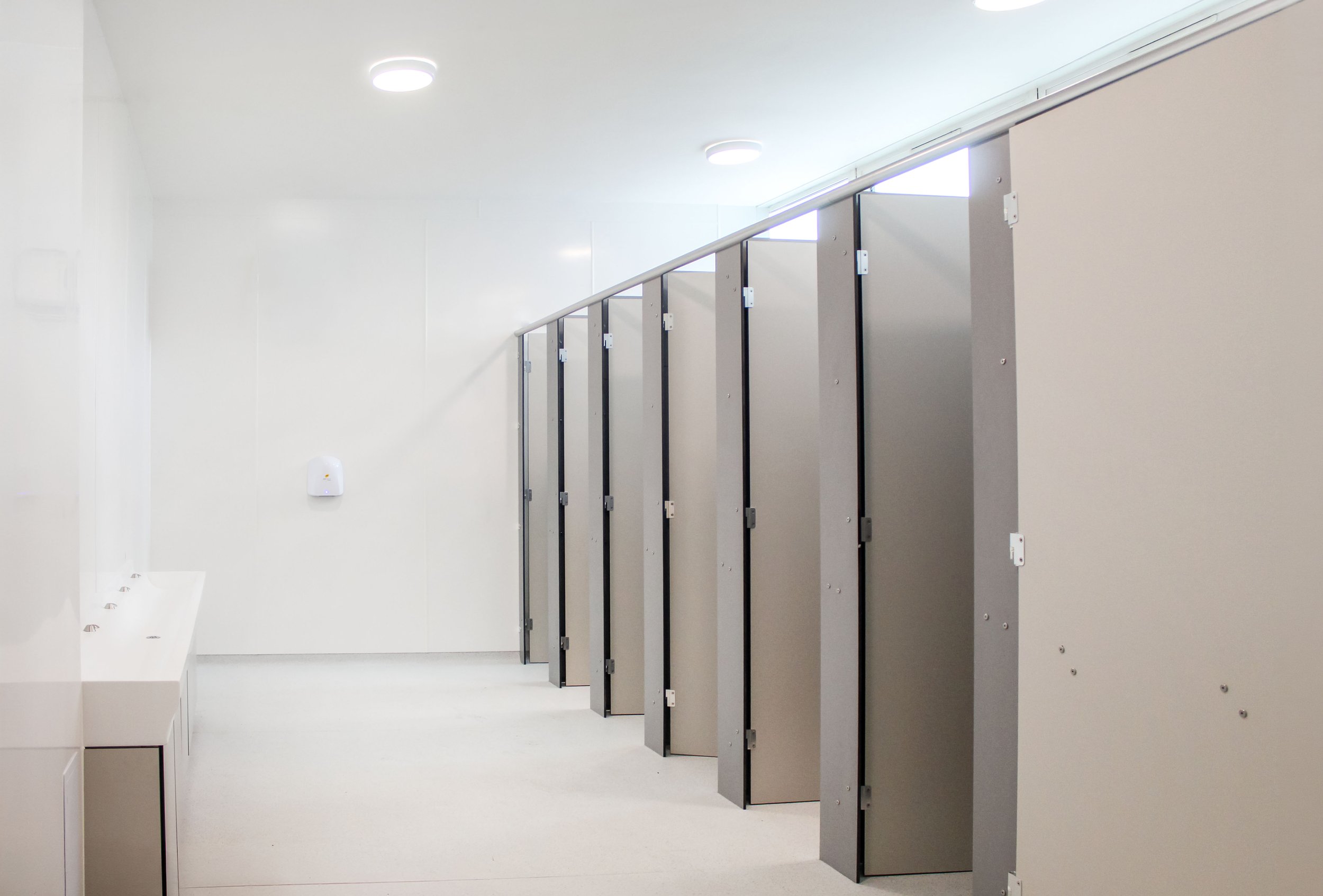 Public restroom with beige stalls, white sinks, and a white wall-mounted hand sanitizer dispenser.