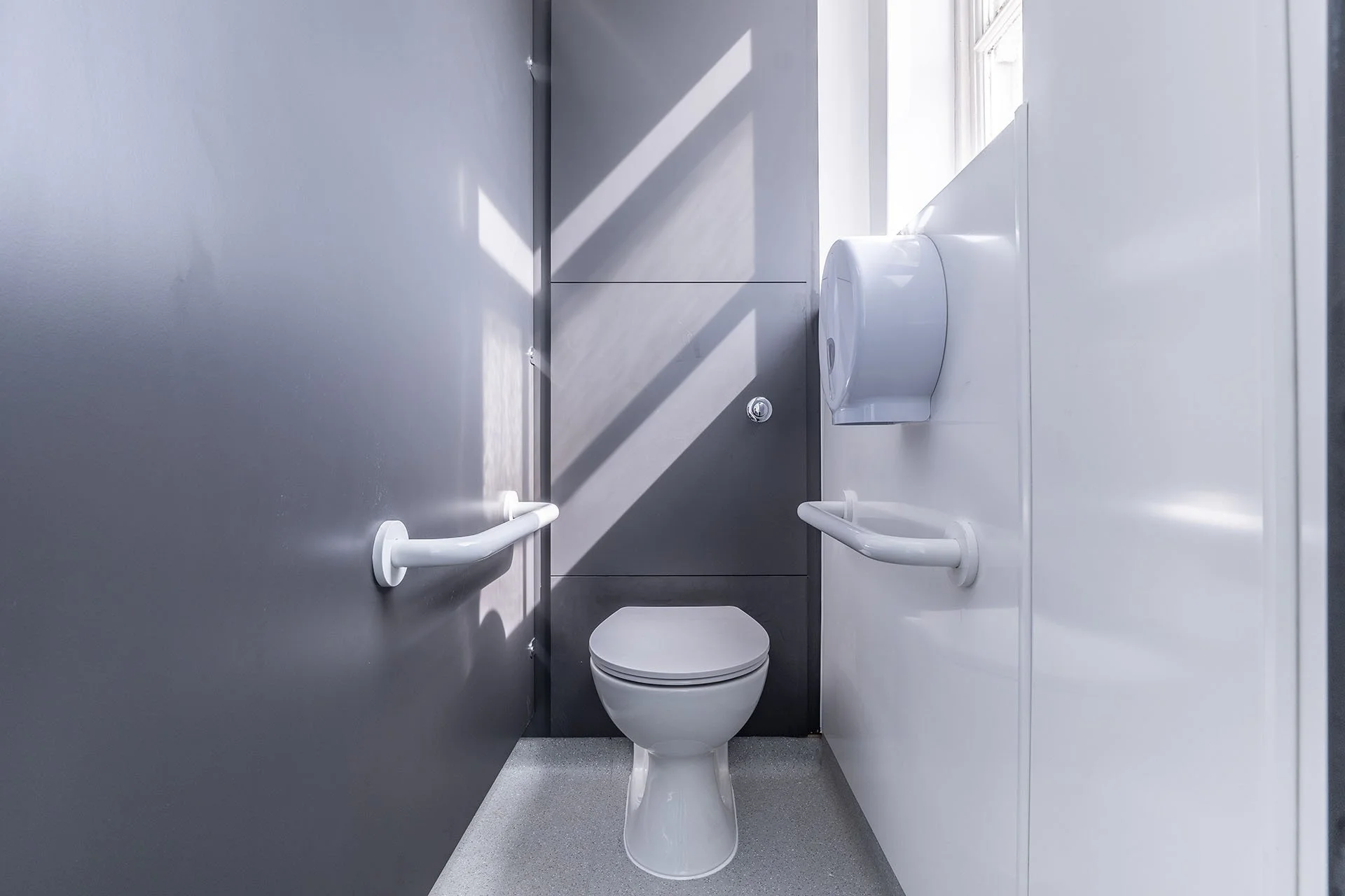 Accessible bathroom with gray and white walls, a toilet, grab bars, and a paper towel dispenser.