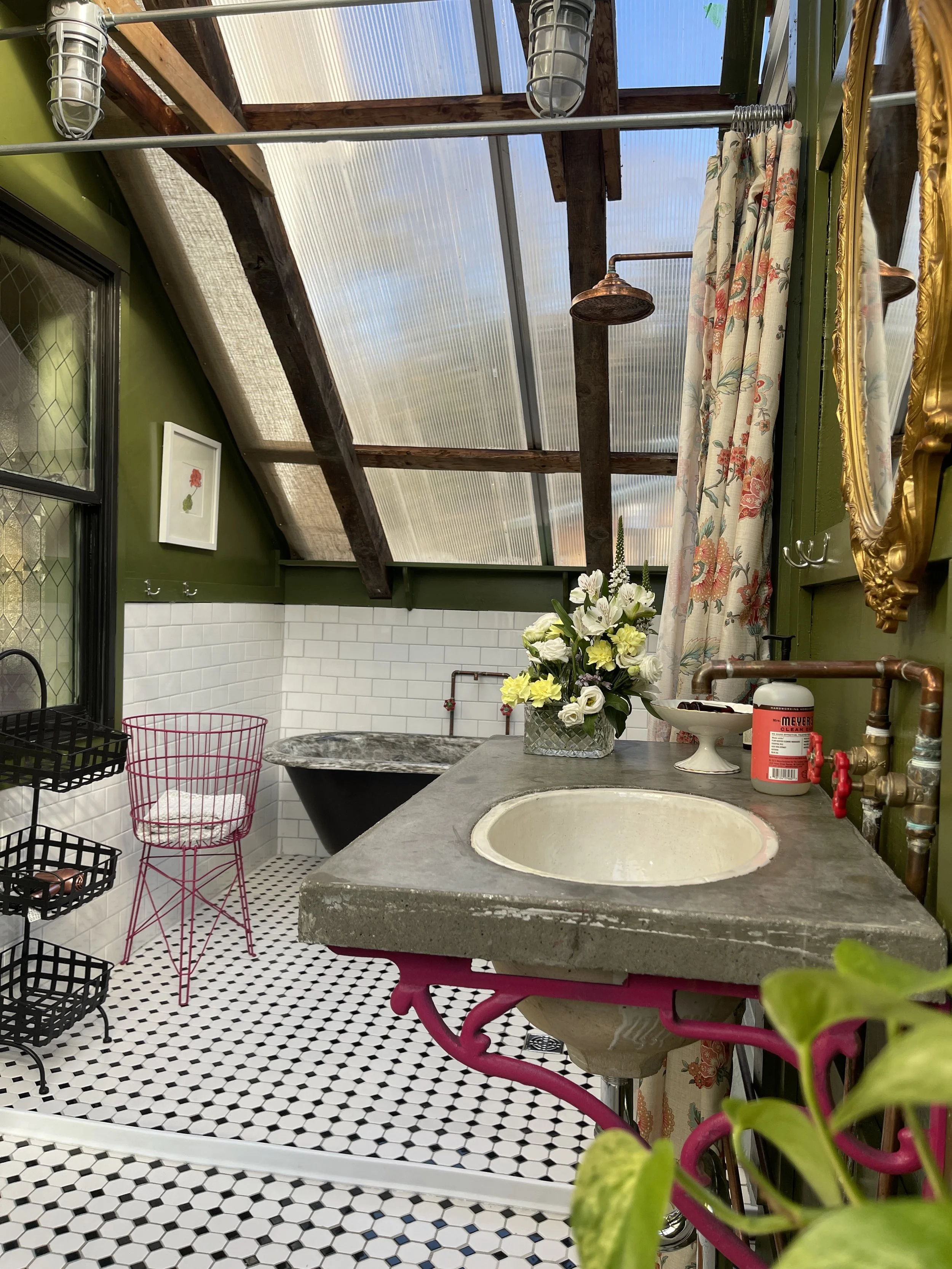 Bathroom with green walls, white subway tile, black bathtub, pink wire chair, black wire shelf, flower arrangement on concrete sink, floral curtain, copper showerhead, skylight, and patterned black and white floor tiles.