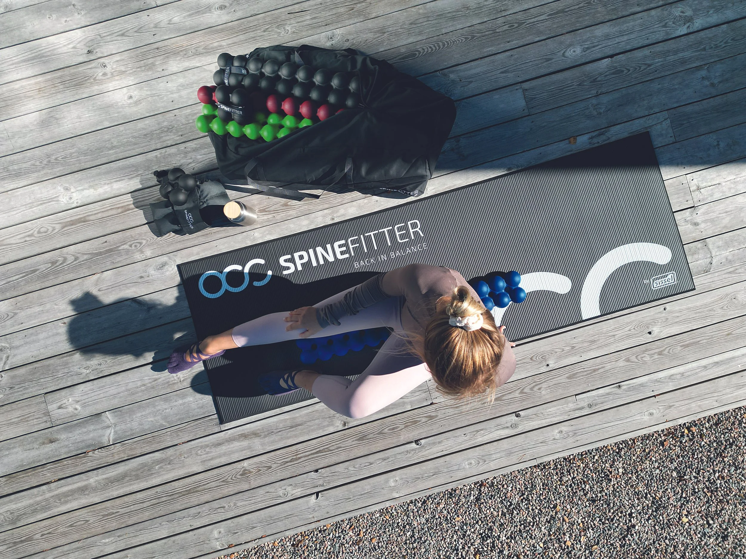 Frau beim Yoga auf einer schwarzen Matte mit SPINEFITTER-Logo, daneben gefüllte Beutel und Wasserflasche auf Holzdeck.