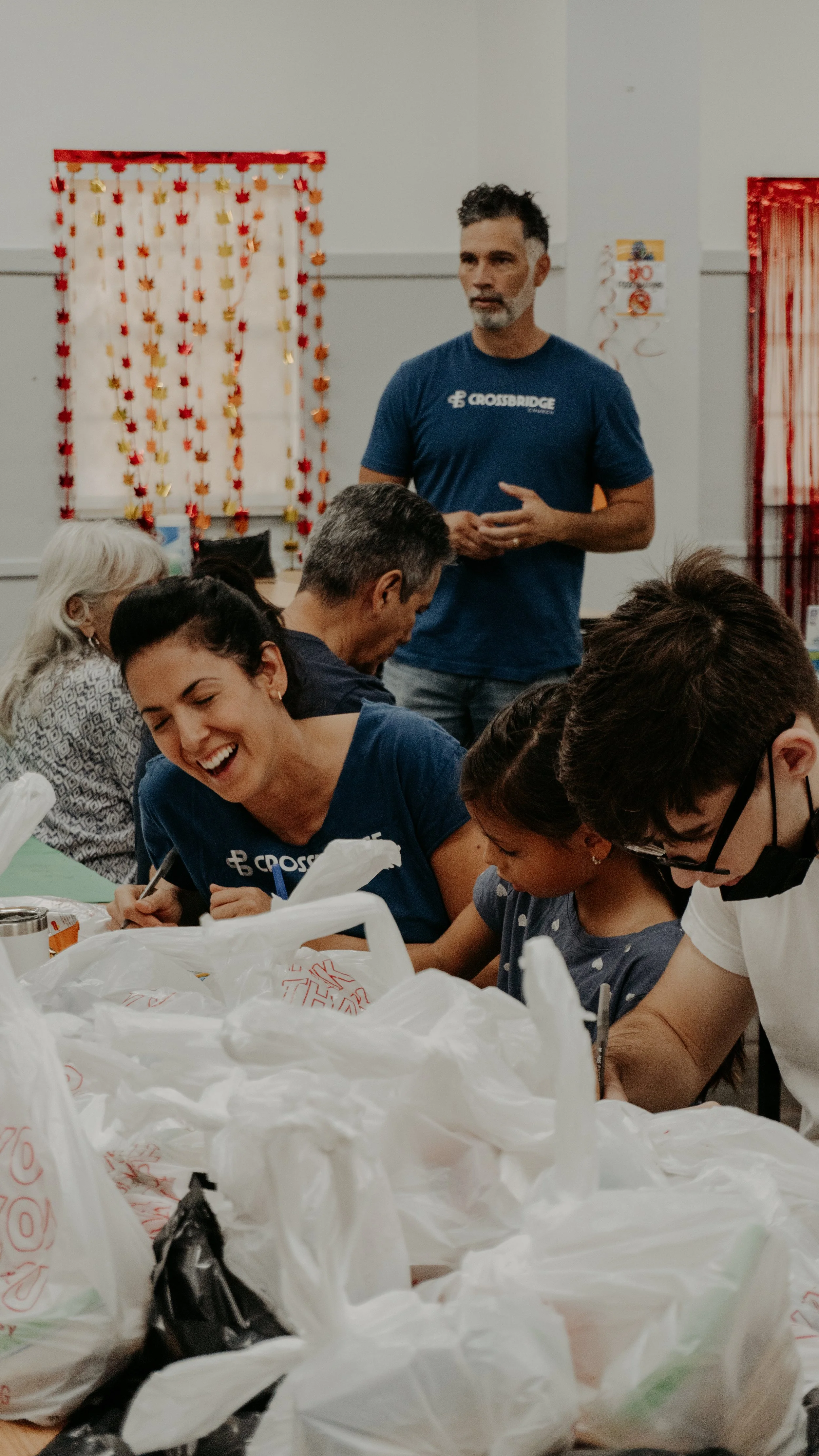 A man in a blue shirt stands in the background, while a woman and others sit at a table, smiling and working with plastic bags and paper. The setting is decorated for a festive event with red, orange, and gold fall-themed decorations on the wall.
