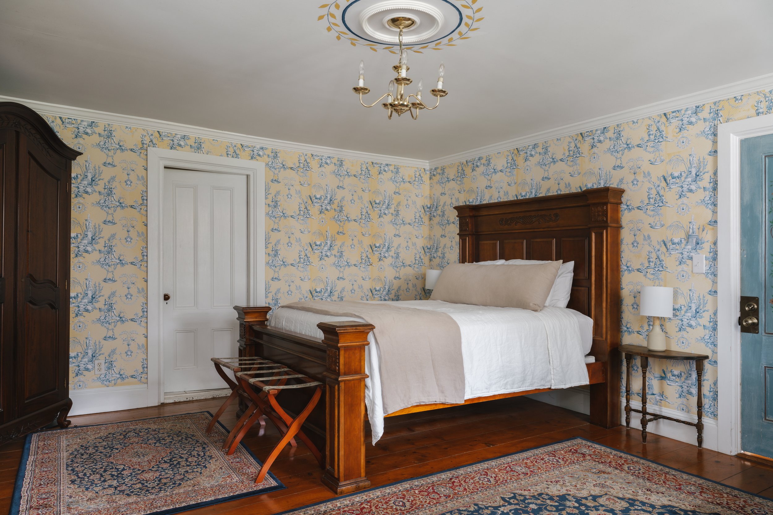 A bedroom with a large wooden bed, a nightstand with a lamp, a blue door, an arched rug, an antique wardrobe, and wallpaper with a pastoral scene pattern.