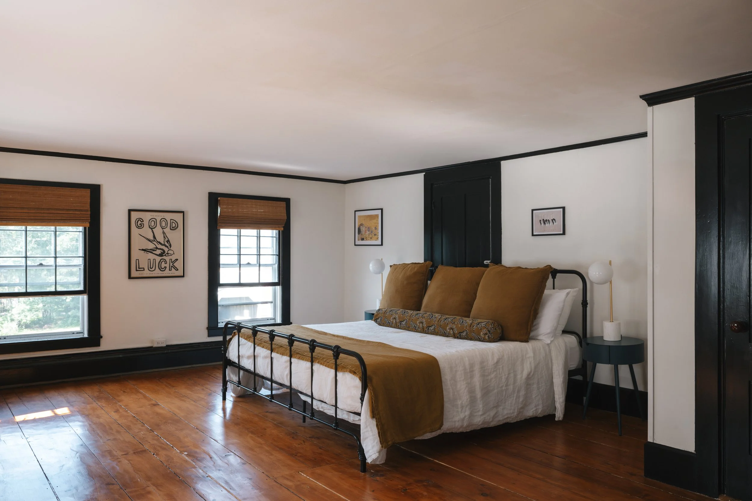 Bedroom with a black metal bed frame, white bedding, large brown pillows, and a decorative pillow, two windows with wooden blinds, wooden floor, black trim, and wall art.