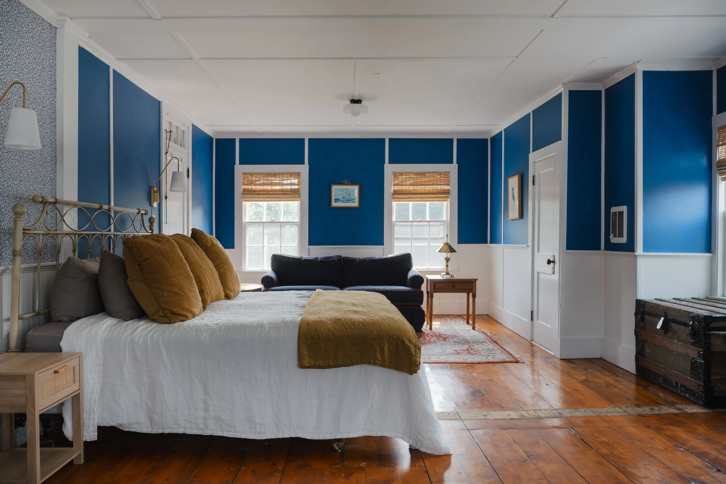 Spacious bedroom with blue and white paneled walls, wooden floors, a bed with white linens and yellow accent pillows, a wooden nightstand, and a sitting area with a sofa and side table.