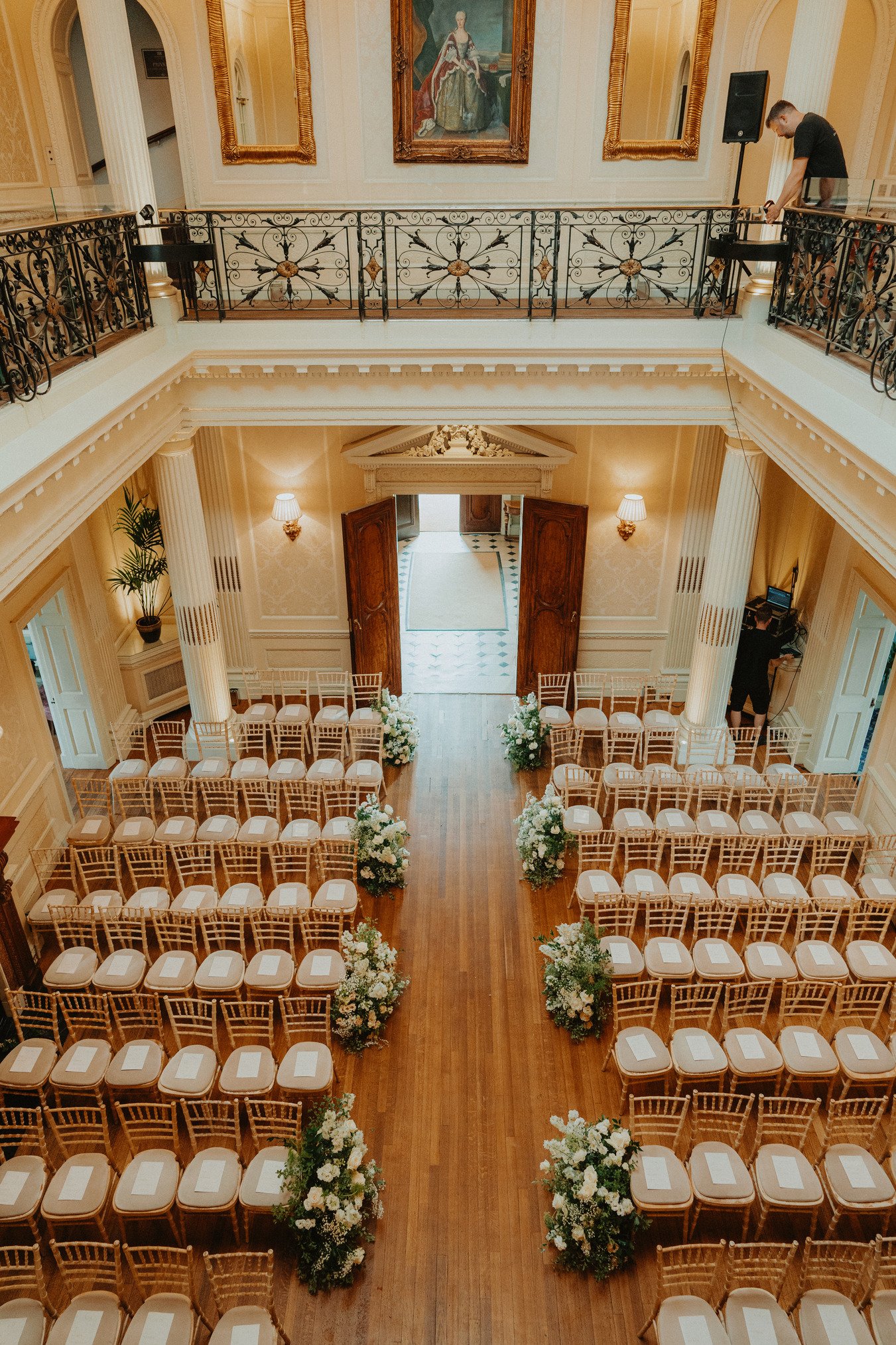 Elegant indoor wedding venue setup with rows of chairs and floral arrangements, ornate balcony, and classical paintings.
