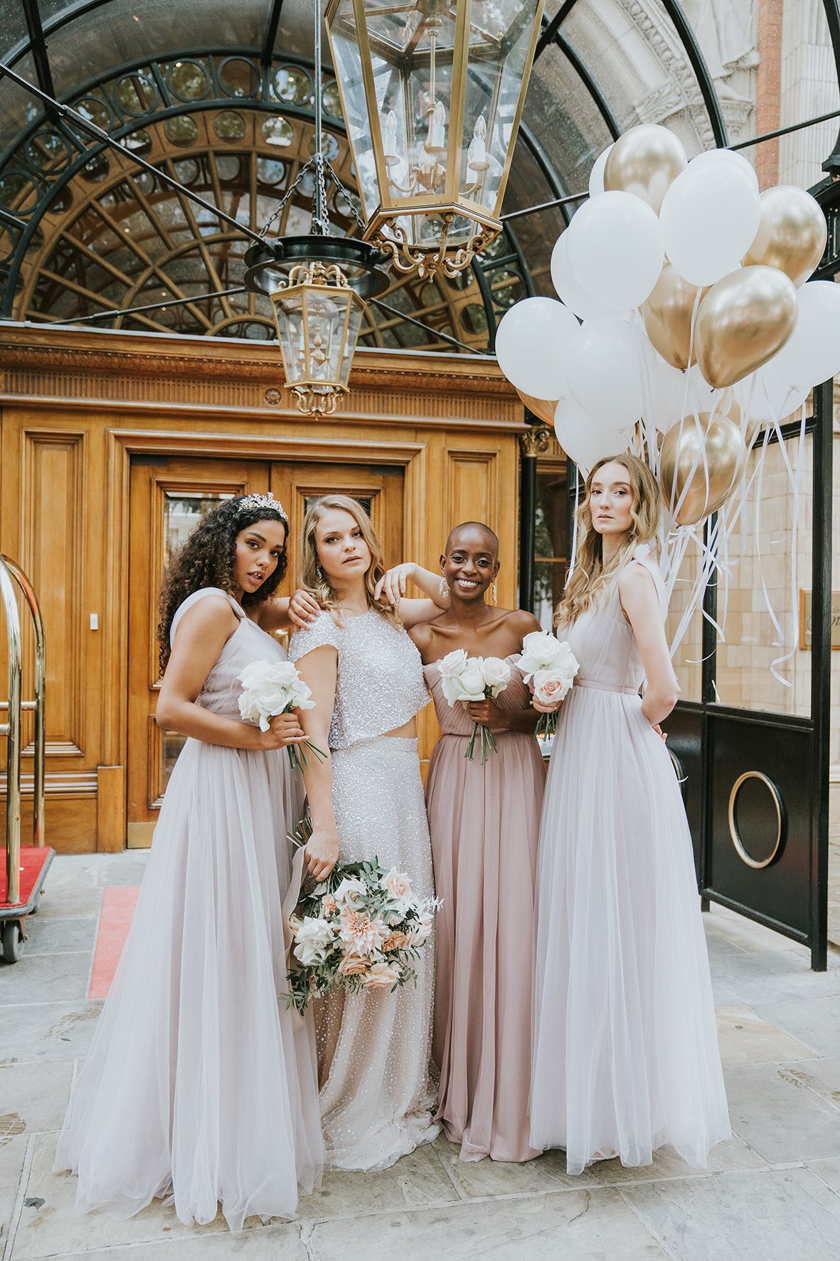 Bride and bridesmaids posing with bouquets, wearing formal dresses, with balloons and a wooden door backdrop.