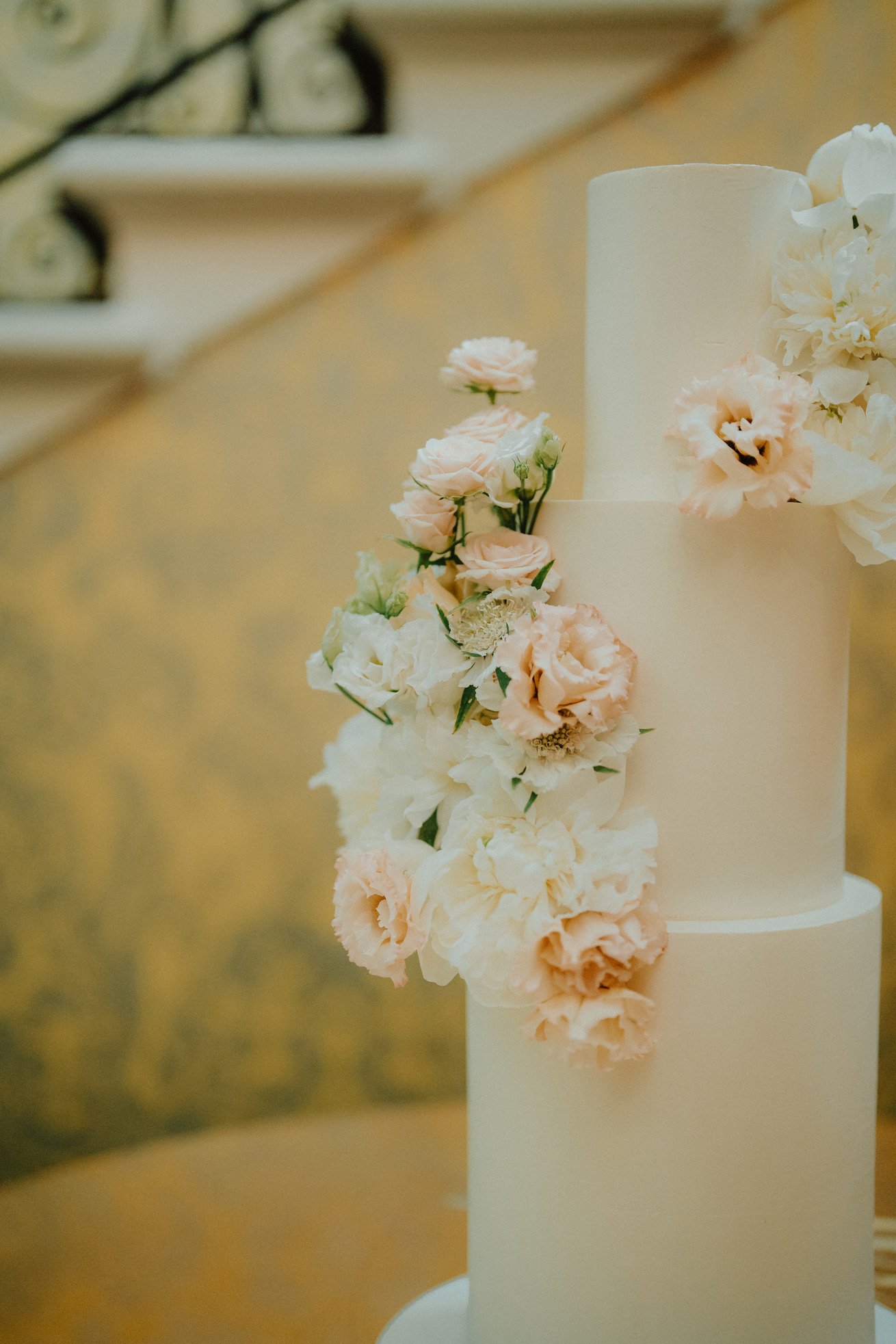 Elegant tiered wedding cake with ivory and blush flowers on staircase background.
