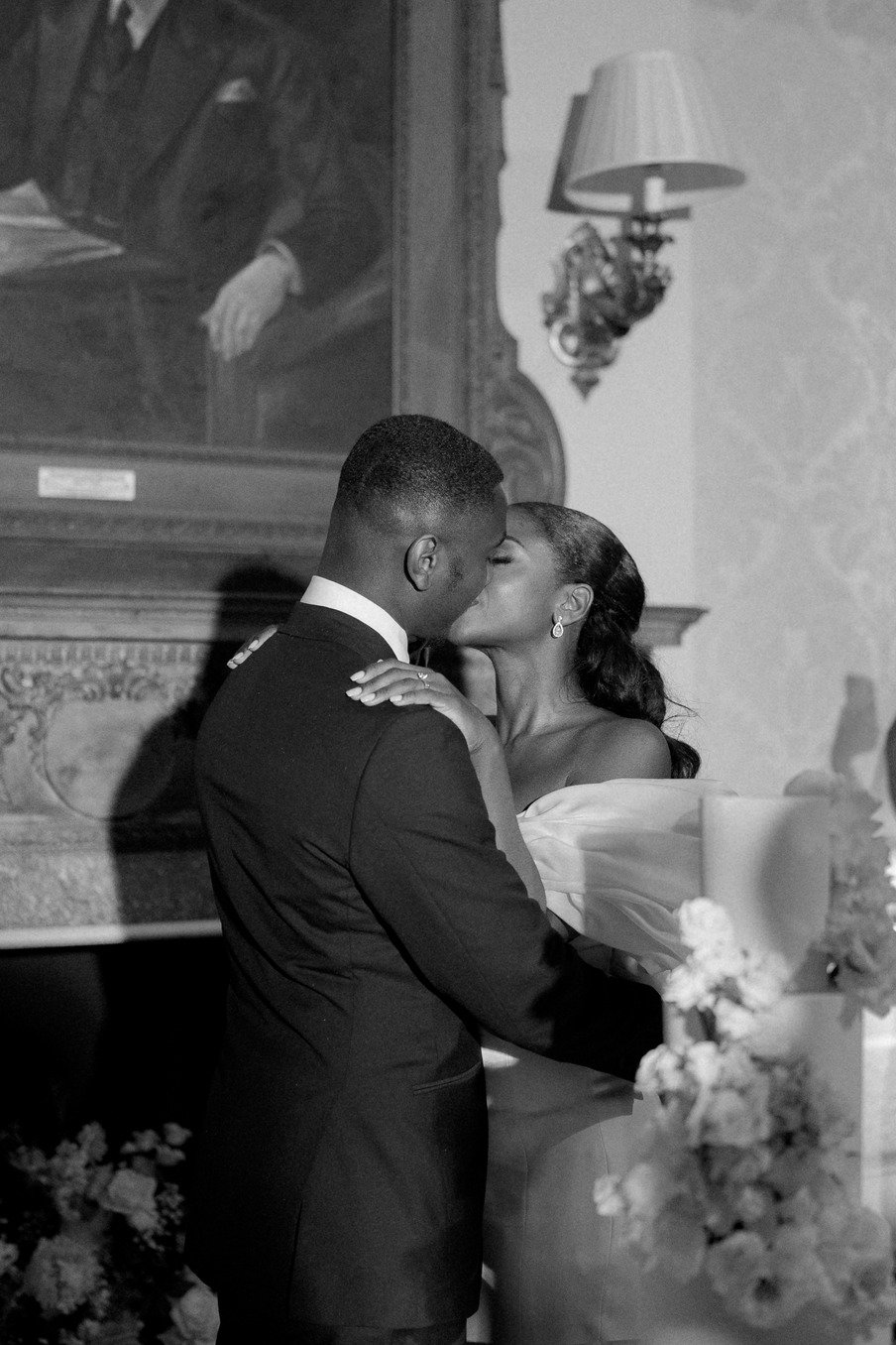 A couple kissing in front of floral decorations and a large portrait in a room with classic decor.