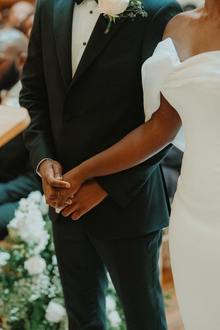 Wedding couple holding hands, groom in black suit with white boutonniere, bride in white gown.