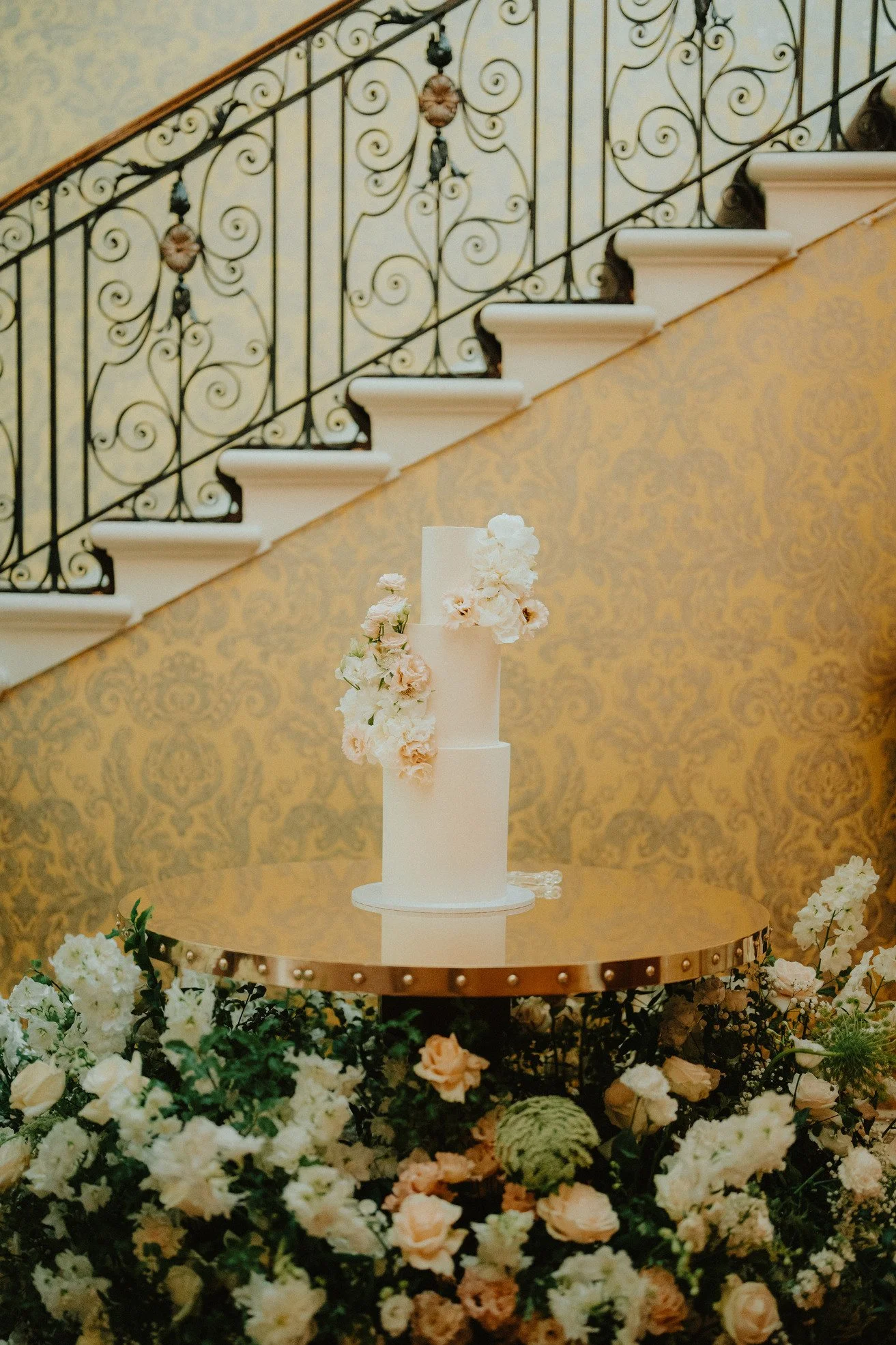 Three-tier white wedding cake with floral decorations on a table, ornate staircase background, surrounded by flowers.