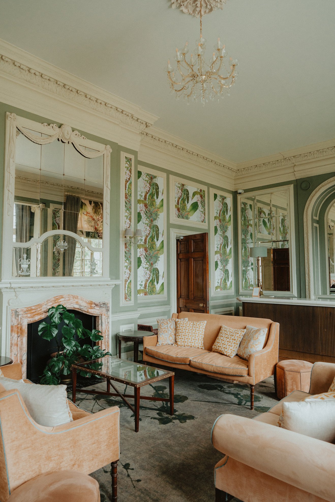 Elegant vintage living room with patterned wallpaper, mirrored walls, chandelier, mustard-colored sofas, glass coffee table, and plants.