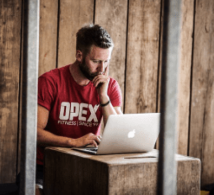 Man with a beard wearing a red OPEX t-shirt working on a laptop at a wooden table in a rustic setting.