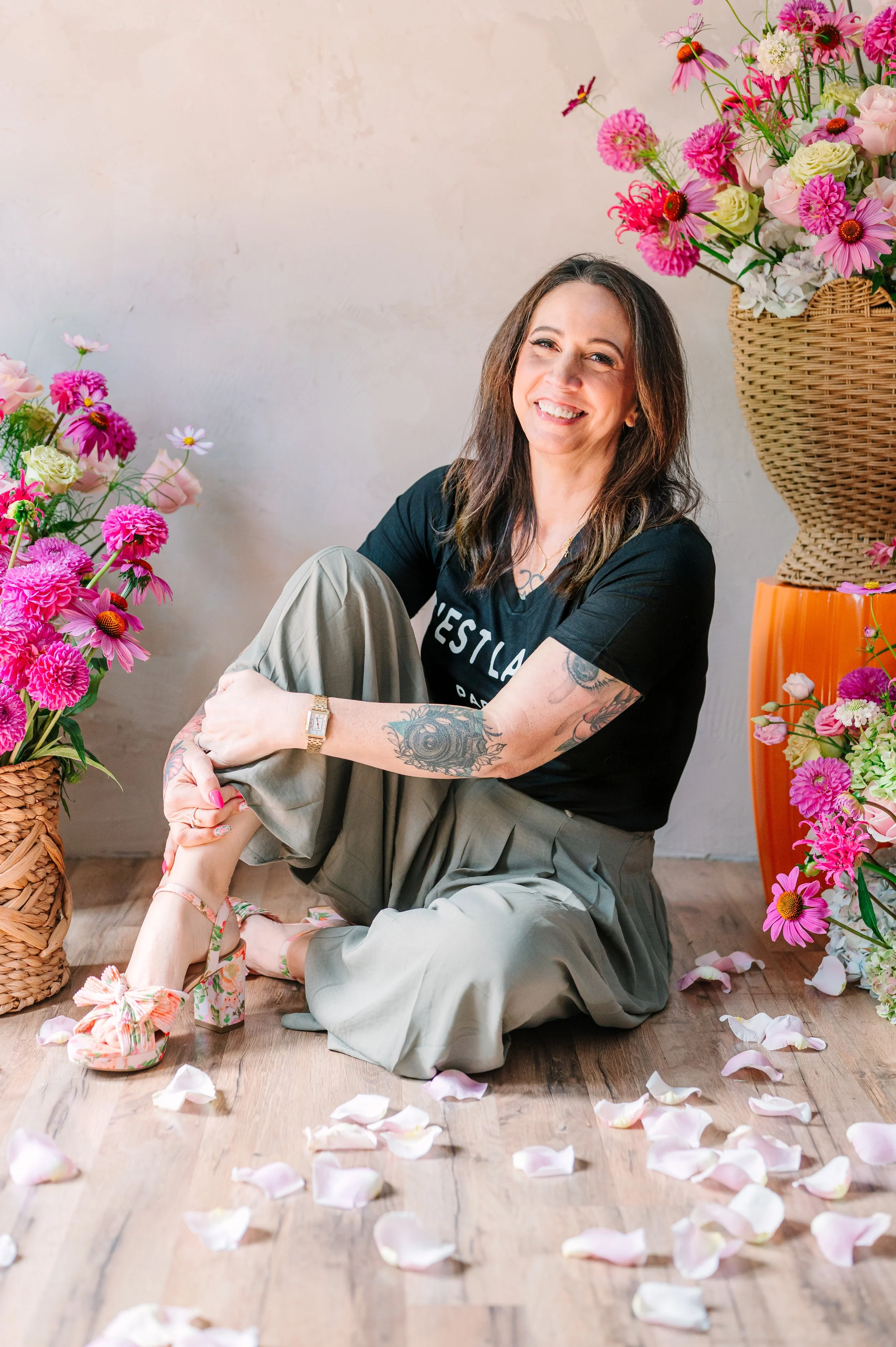 A woman with tattoos sitting on a wooden floor, surrounded by pink and white flowers, smiling at the camera. She is wearing a black t-shirt and light-colored pants, with floral high heels, flower petals scattered around her.