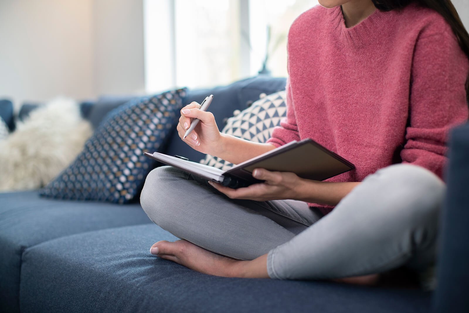A woman sits cross-legged on a couch, writing thoughtfully in a journal in a warm, comfortable home setting. This kind of inward reflection is a natural starting point, but in-person therapy in St. Louis, MO
