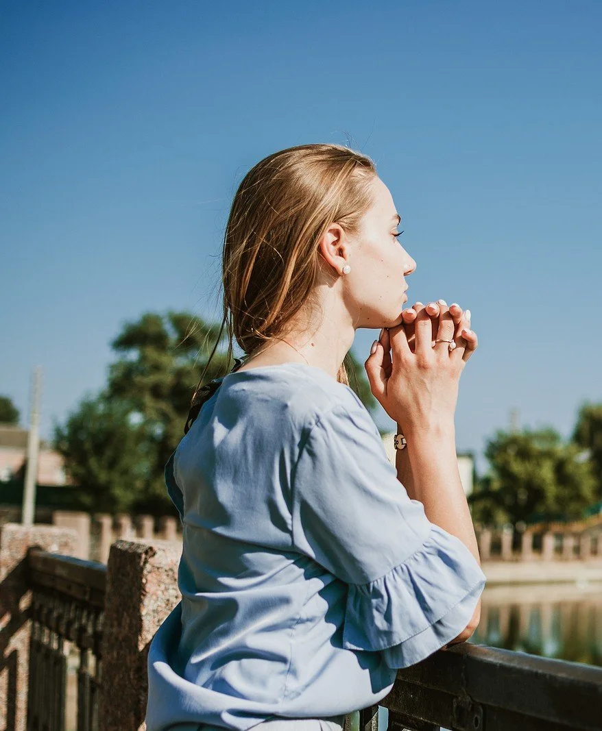 A woman stands outdoors in bright sunlight with her hands clasped under her chin. Learn how IFS therapy in St. Louis, MO can offer support with inward searching. Contact an IFS counselor St. Louis today.