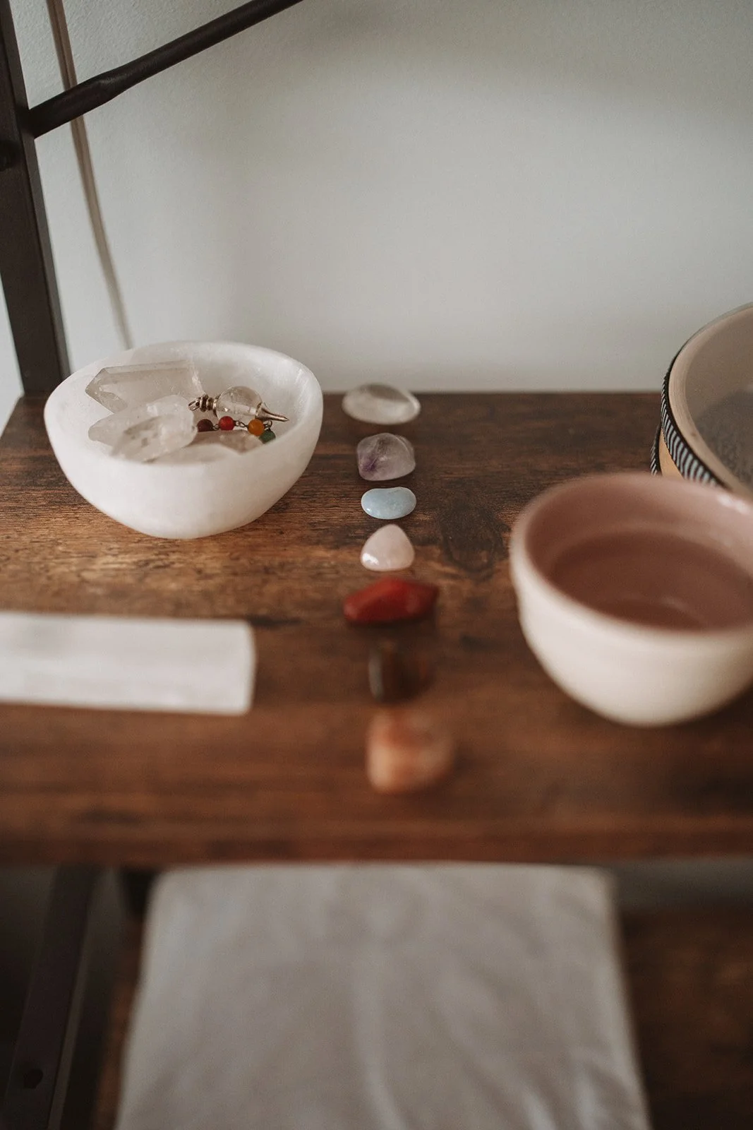 A wooden table with a white shell-shaped bowl containing jewelry, colorful crystals lined up along the top of the table, a white cup with a light pink interior, and a partially visible dish on the right. A white cloth is on the lower part of the table.