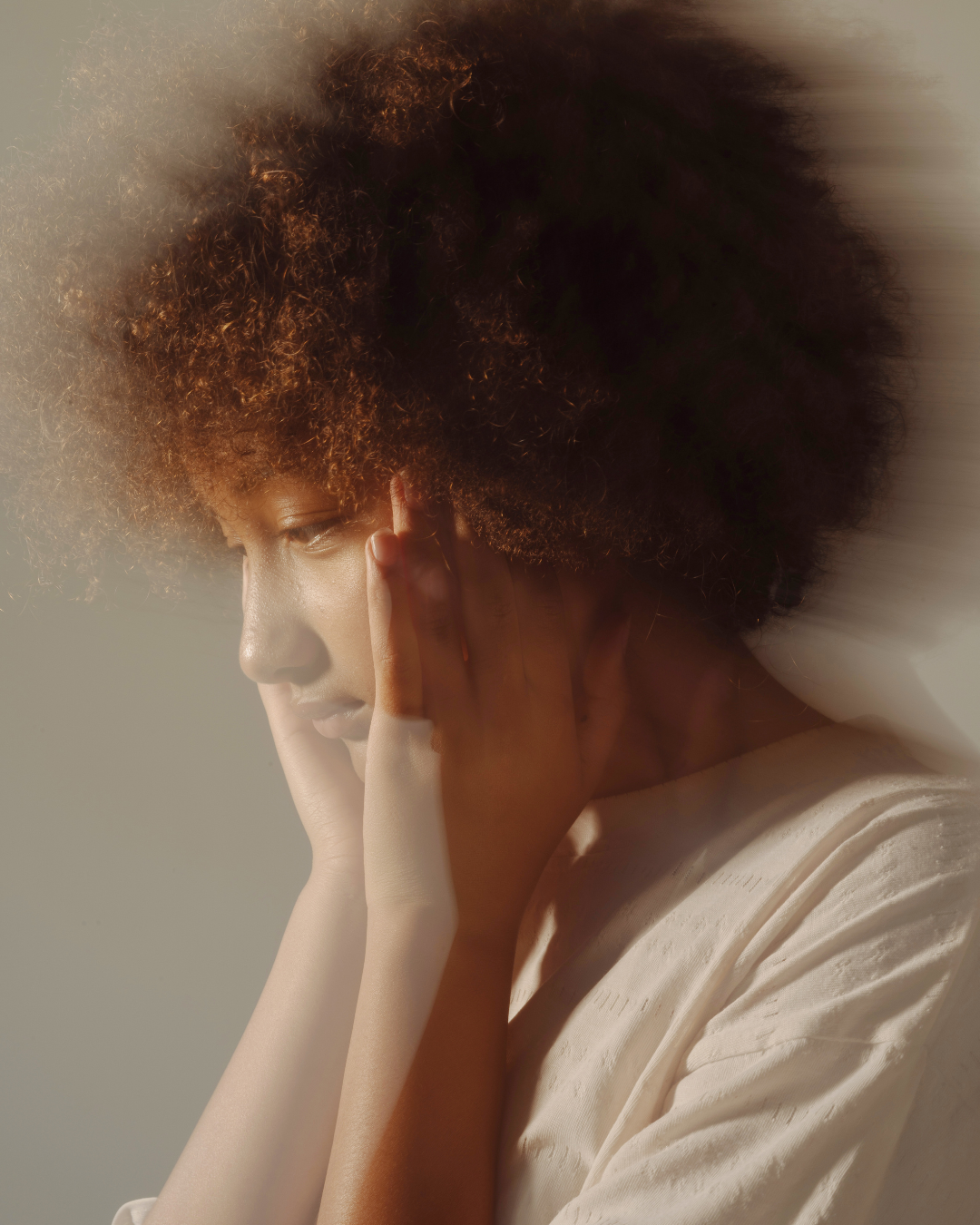 A woman with curly afro hair touching her face, looking downward in a soft light.