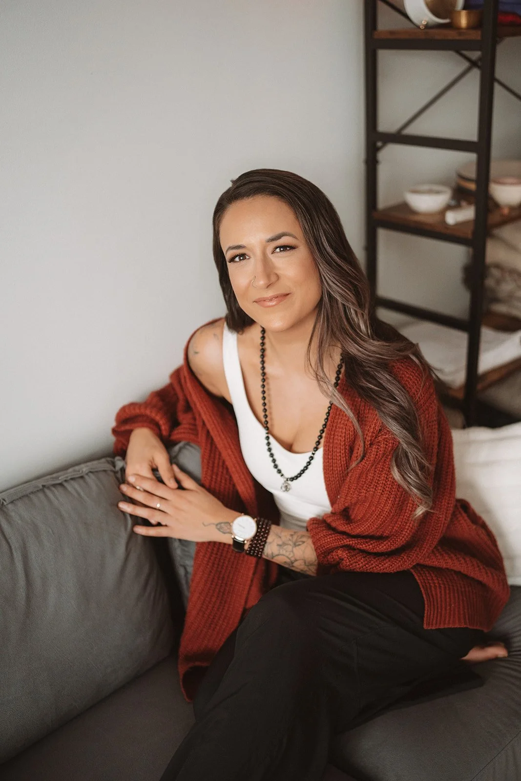 A woman with long brown hair, wearing a white tank top, black beaded necklace, and red cardigan, sitting on a gray couch with a tattooed arm, smiling at the camera.
