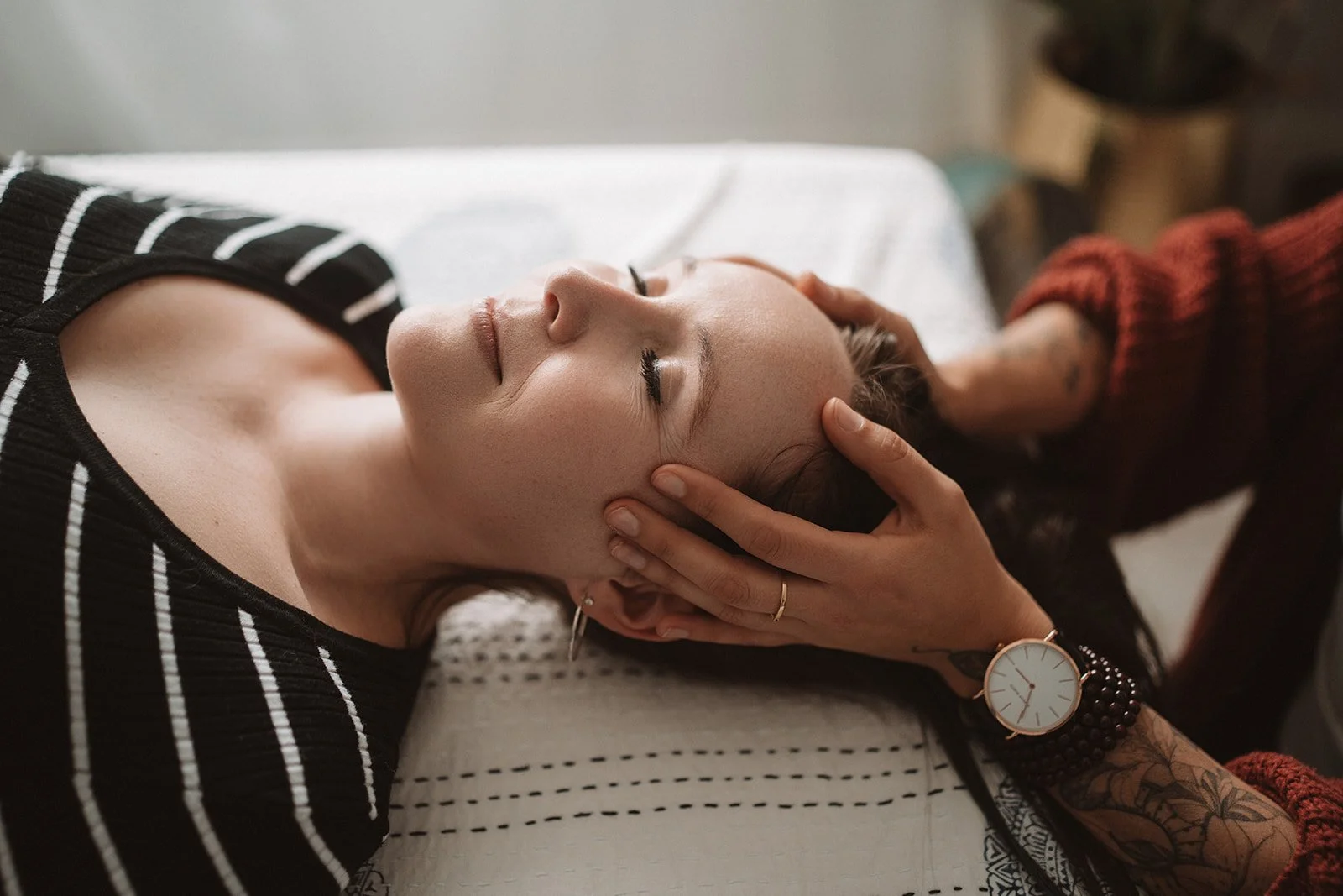 A woman lying on her back with eyes closed, receiving a head massage or facial treatment from another person whose hand is gently on her forehead. The woman has earrings, a tattoo on her arm, and is wearing a striped shirt and a watch.