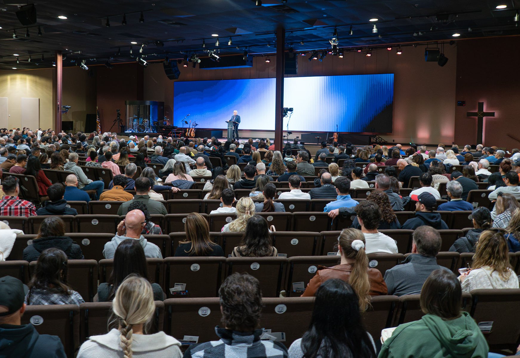 A large crowd gathered inside a church auditorium attending a presentation or service on stage. The stage has a speaker at the podium, with musical instruments and large screens in the background. A cross is visible on the right wall.