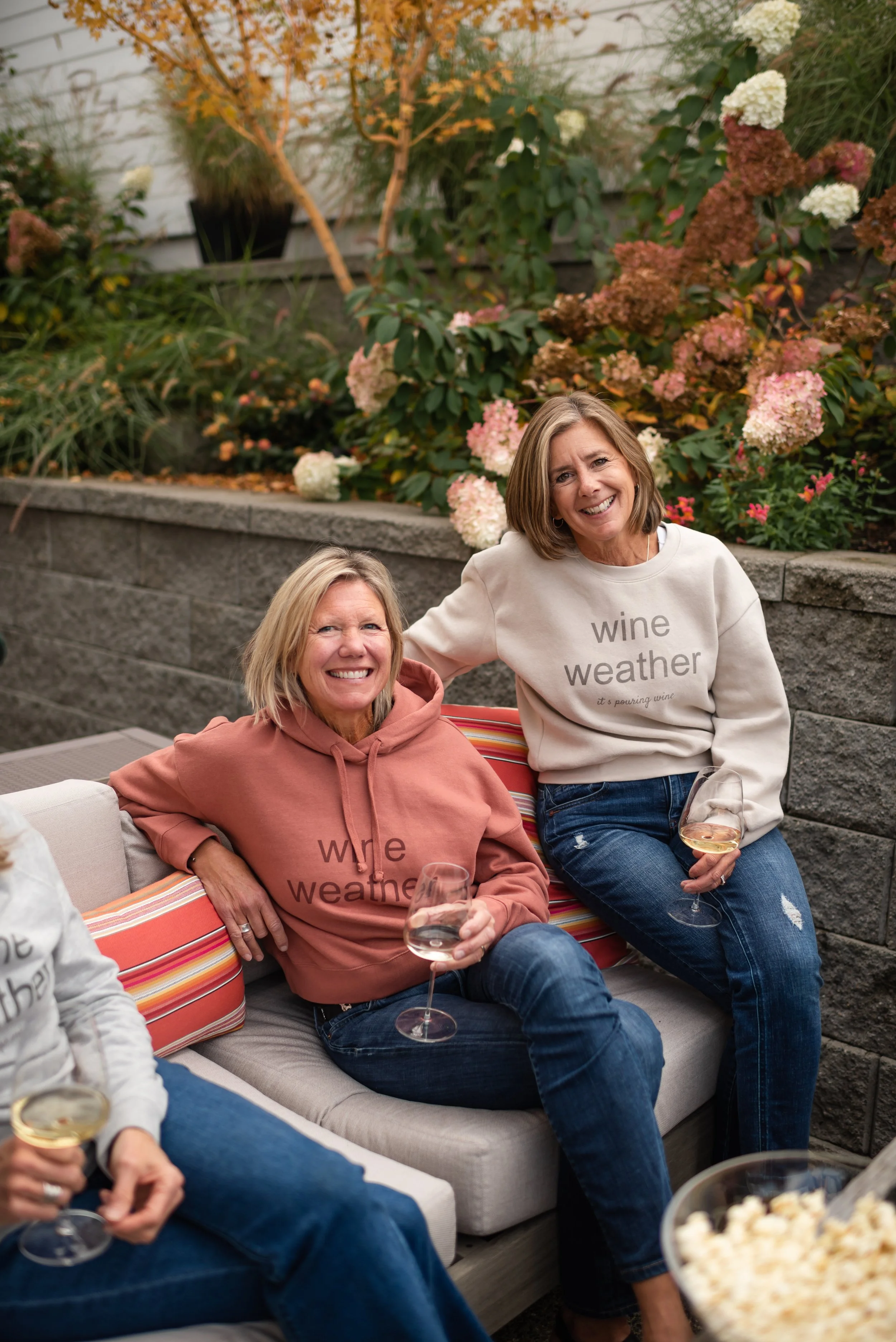 Two women sitting on an outdoor sofa, smiling, wearing matching 'wine weather' hoodies, holding wine glasses, in a garden with flowers and plants in the background.