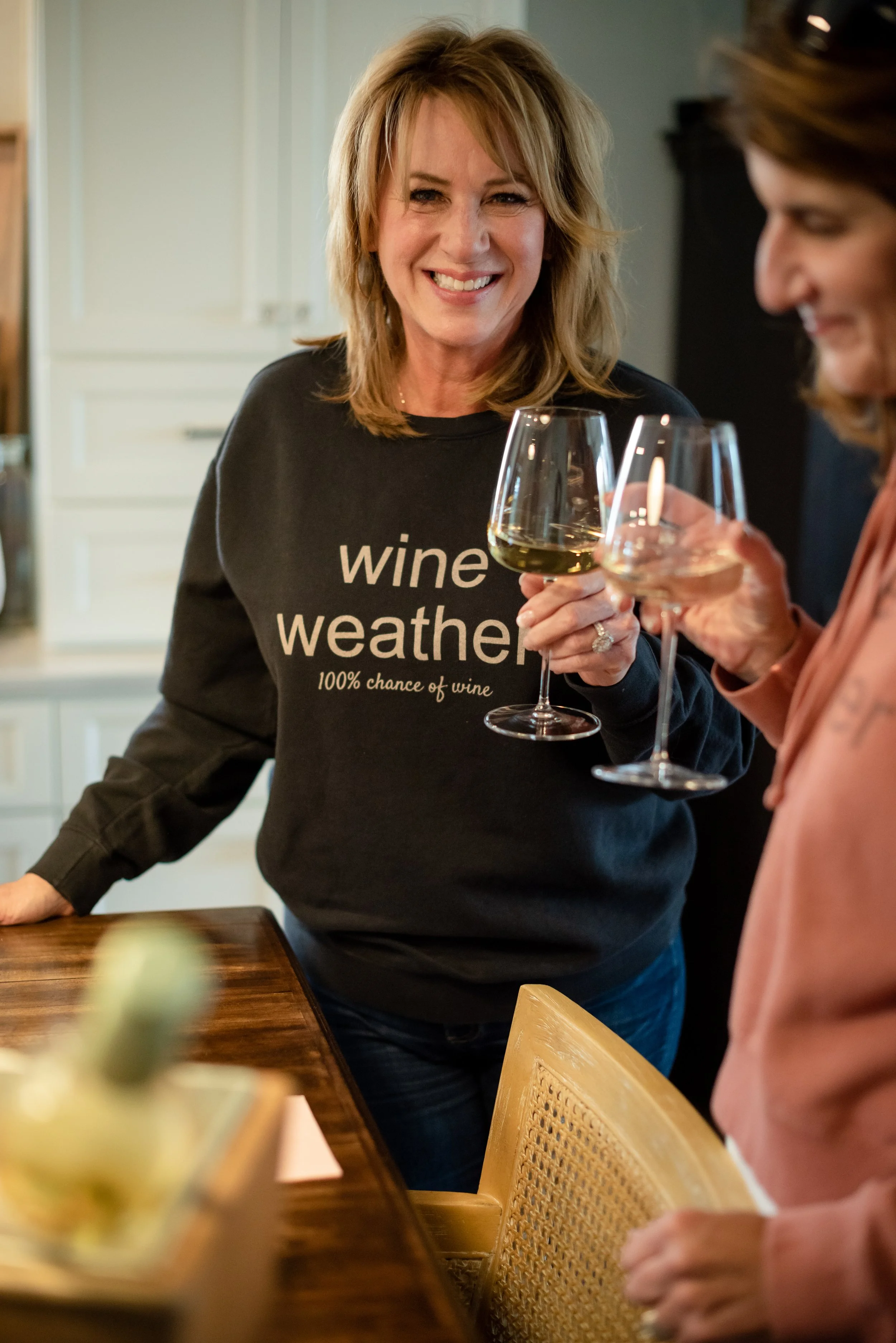 Two women holding wine glasses, smiling and conversing in a home kitchen or dining area. One woman is wearing a black sweatshirt with the text "wine weather" and a subtitle "100% chance of wine".