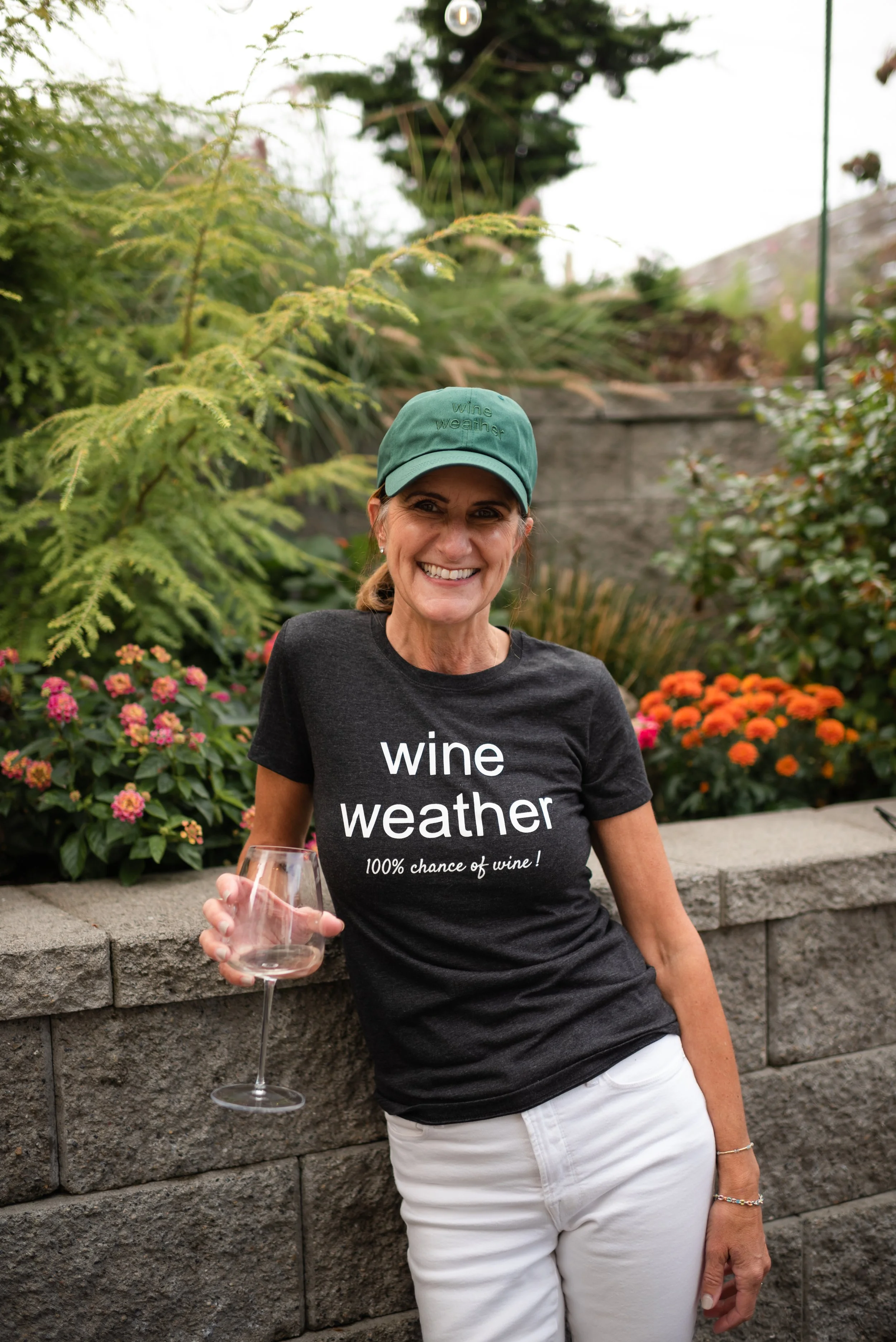 A smiling woman wearing a green cap and a black t-shirt with the text "wine weather 100% chance of wine!" holding a glass of wine, standing outdoors in front of a garden with colorful flowers and greenery.